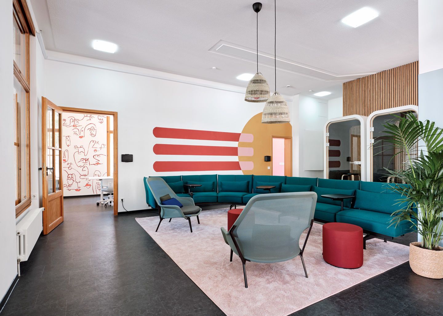 Helle Bürolounge mit türkisfarbenen Sofas, zwei Vitra Slow Chairs Sesseln und roten Hockern auf einem rosa Teppich.