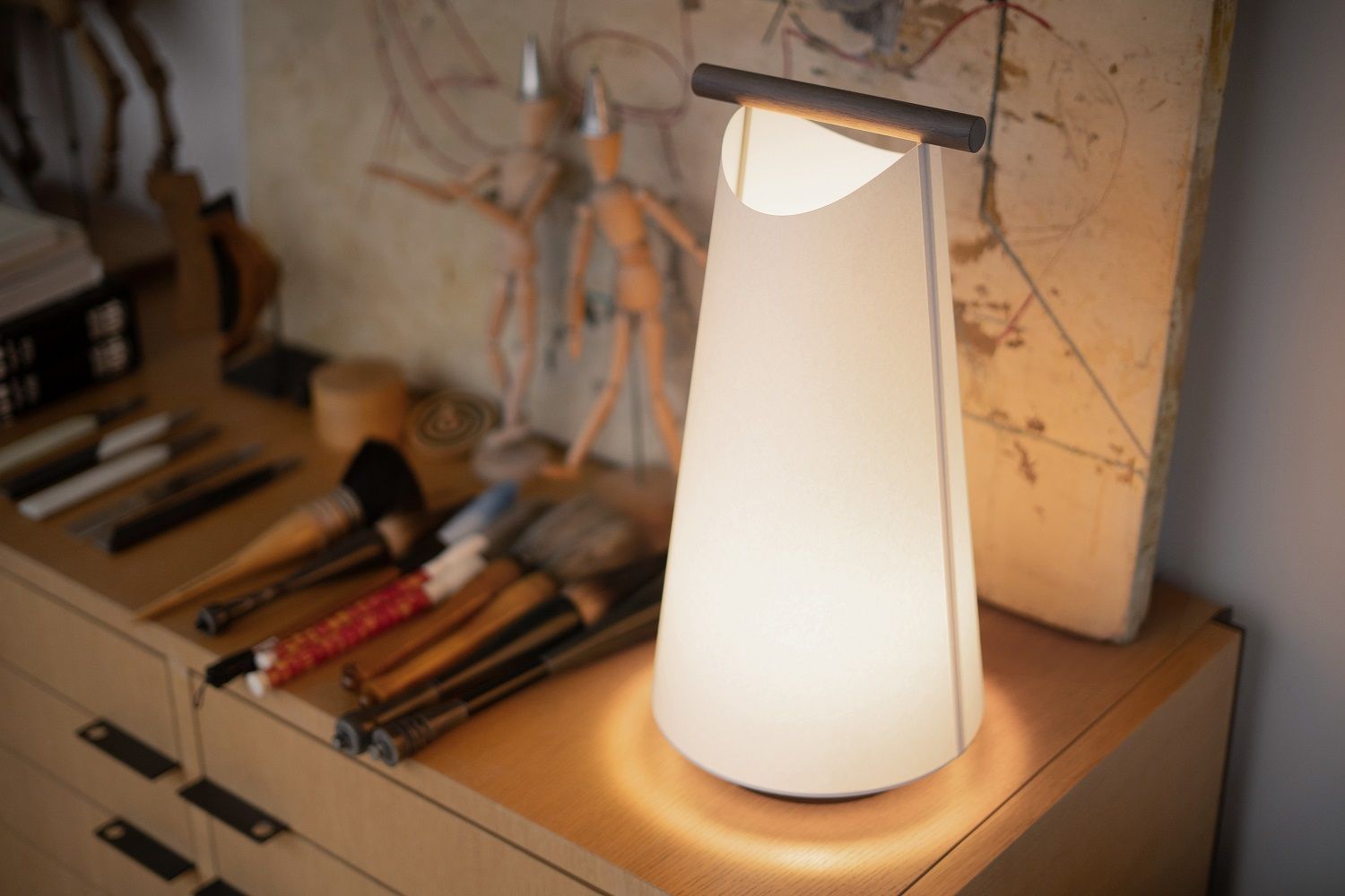 Nomon Catay Tischleuchte, moderne Akku-Tischlampe mit kegelförmigem Lampenschirm und Holzdetail auf einem Schreibtisch.