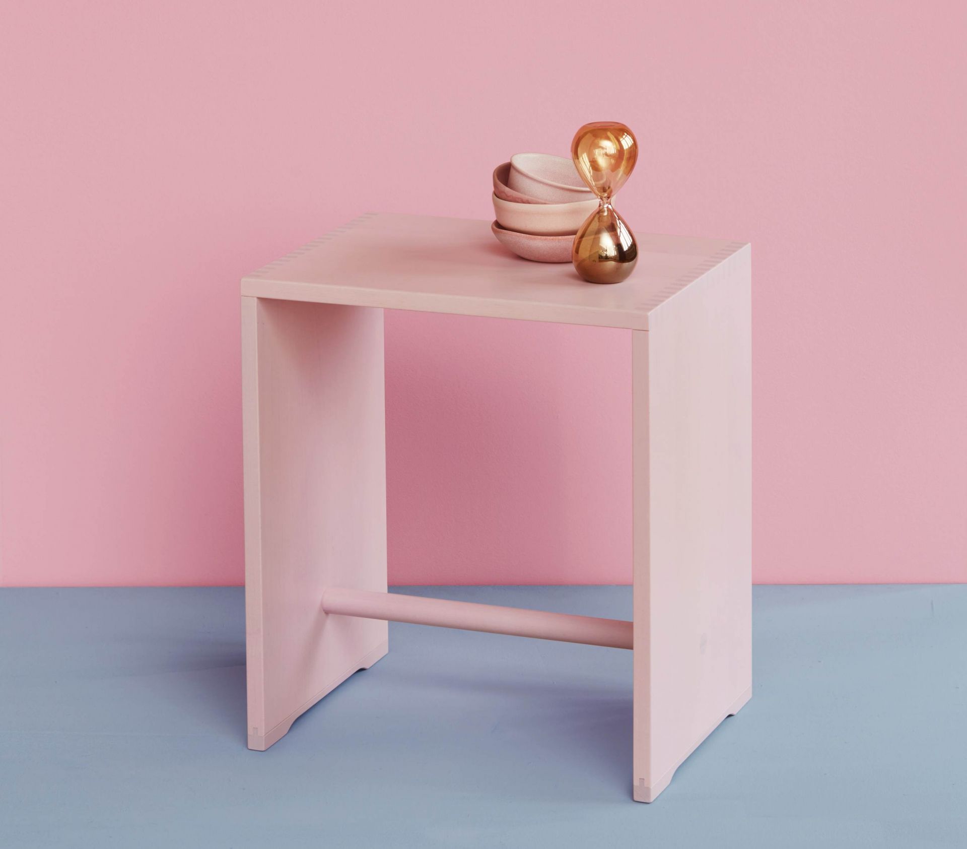 Rosa Hocker aus Holz mit Schalen und goldener Sanduhr vor rosa und blauem Hintergrund.