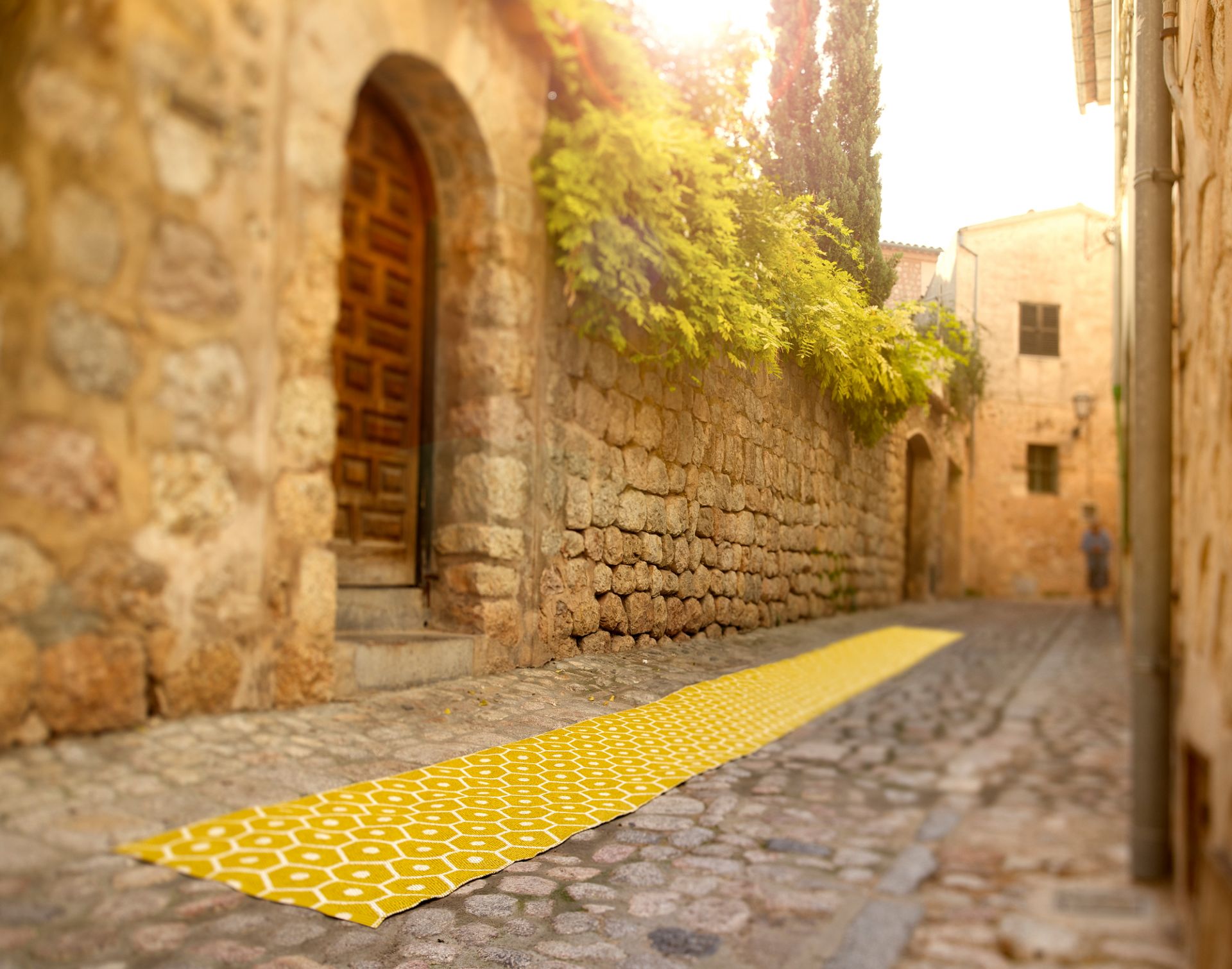 Pappelina Honey Kunststoffteppich 70x225 cm auf Kopfsteinpflaster in Gasse. Gelber Teppich mit geometrischem Muster.