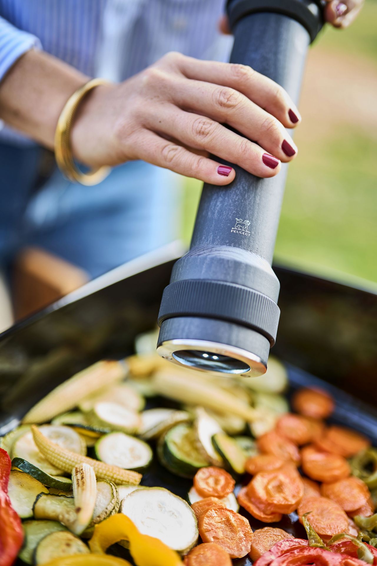 Peugeot BBQ Pfeffermühle in Aktion: Würzen von gegrilltem Gemüse mit graphitfarbener Mühle im Freien.