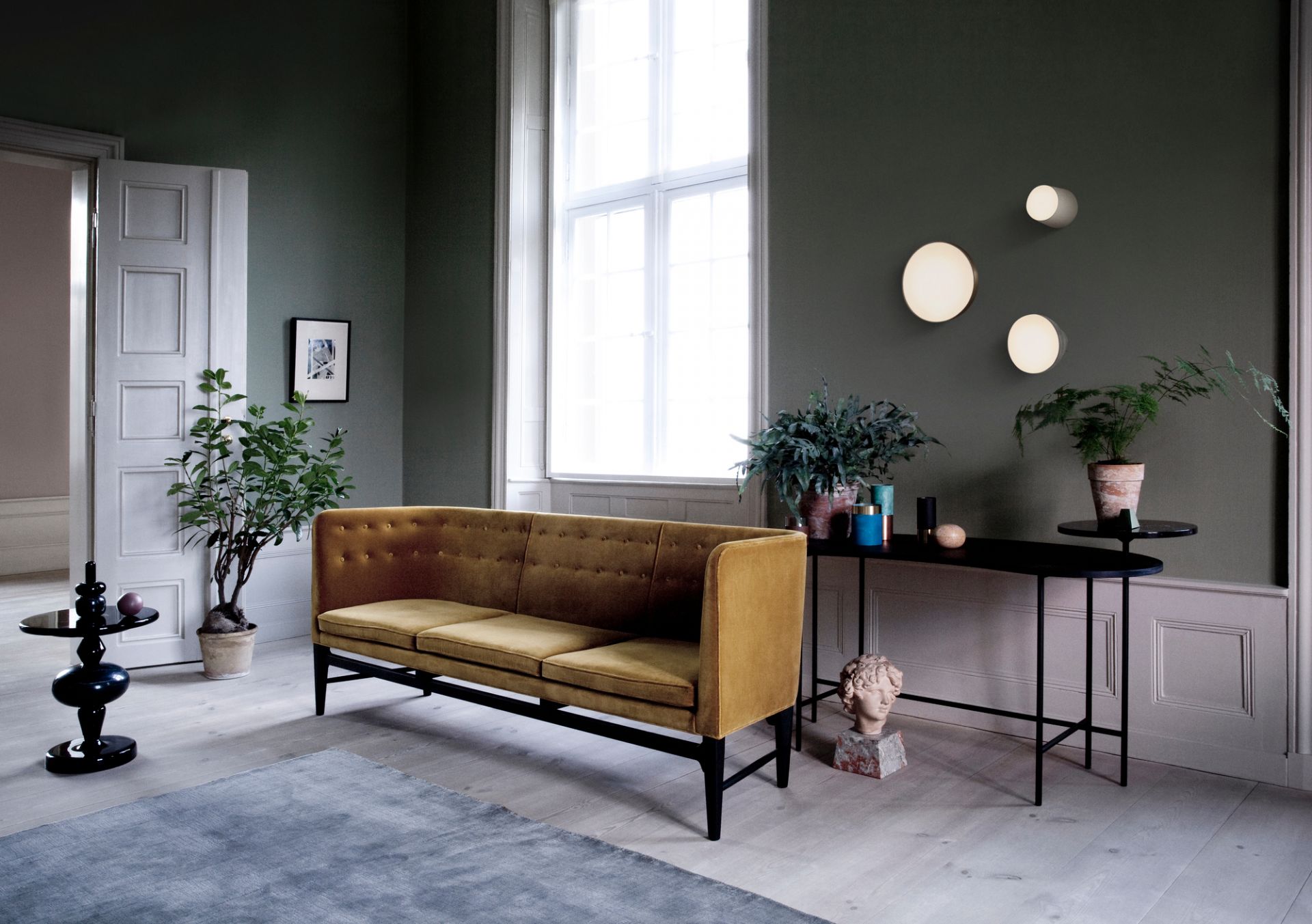 AndTradition Mayor Sofa, 3-Sitzer, goldener Samtbezug, schwarzes Gestell in stilvollem Wohnzimmer mit Pflanzen.