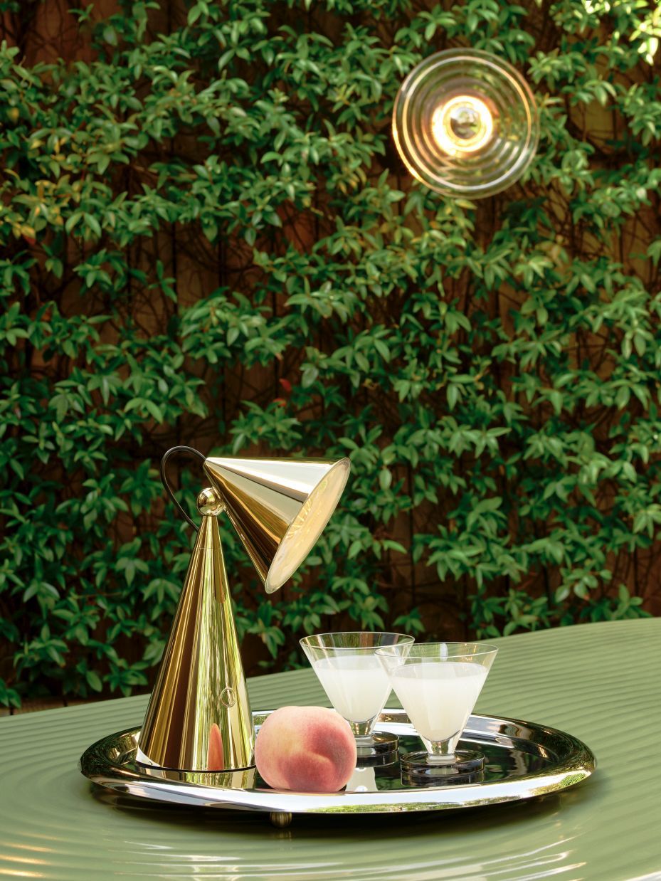 Goldene Tom Dixon Tischleuchte auf Tablett mit Getränken und Pfirsich, im Hintergrund grüne Pflanzenwand und Wandleuchte.