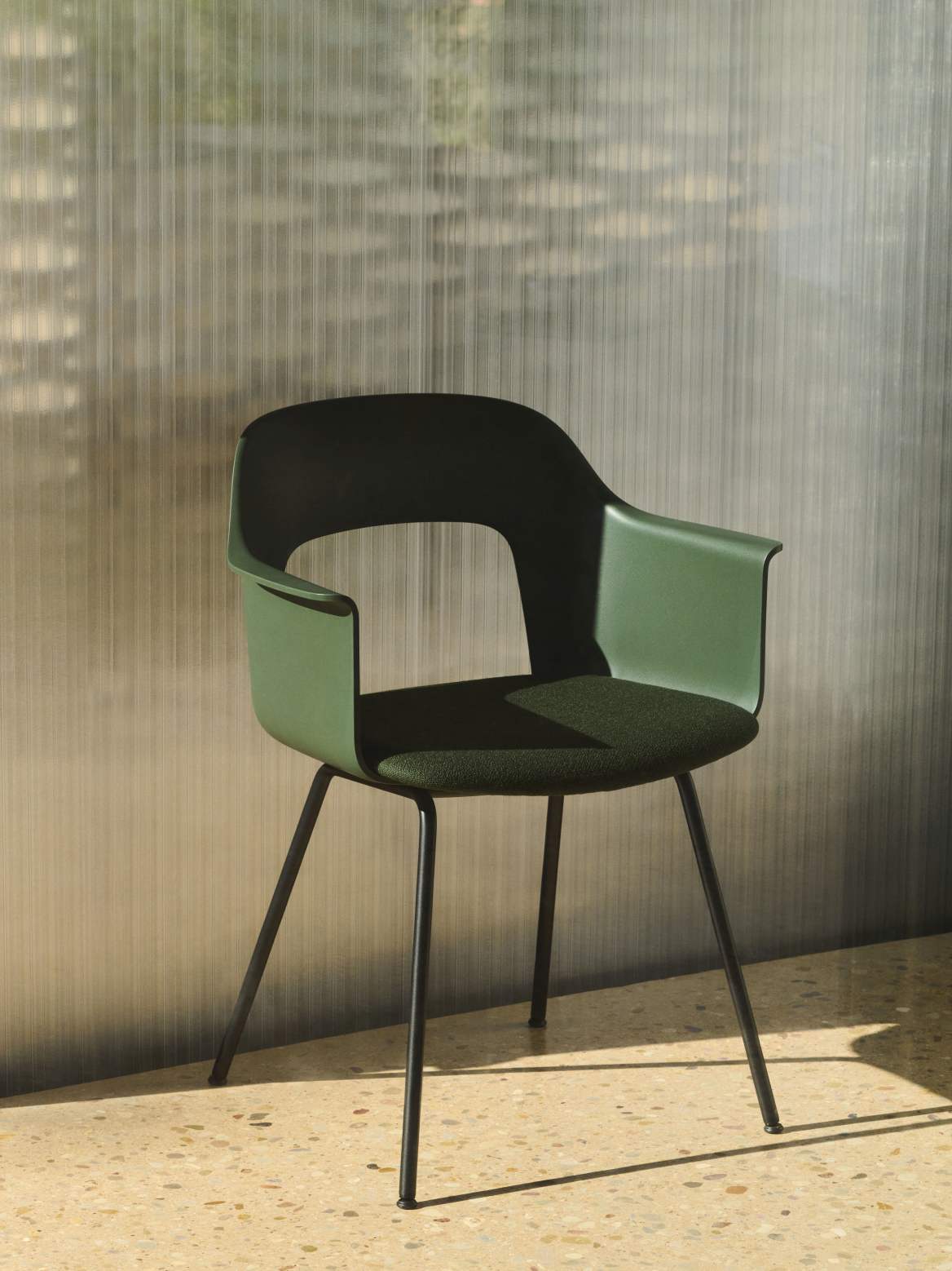 Grüner Hay Armlehnstuhl Layout Armchair 213 mit schwarzem Metallgestell und Stoffbezug vor geriffelter Wand.
