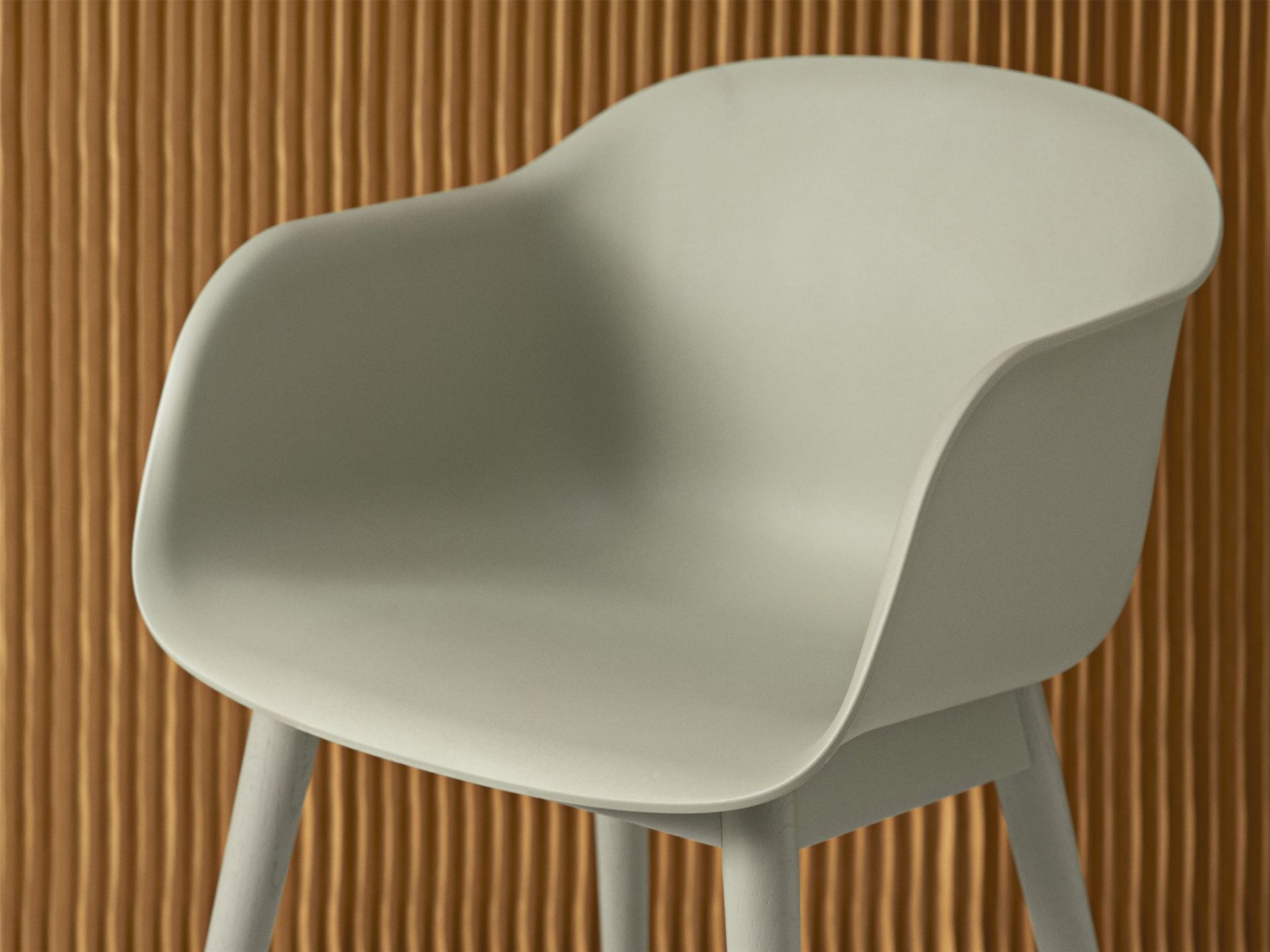 Nahaufnahme des Muuto Fiber Armchair Stuhls mit Holzgestell in sanftem Grün vor geriffelter Wand.