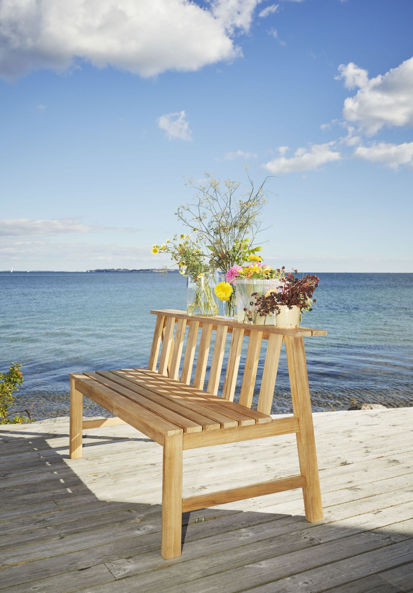 Cadence Teak Bank von Skagerak by Fritz Hansen auf Holzterrasse mit Meerblick und Blumen.