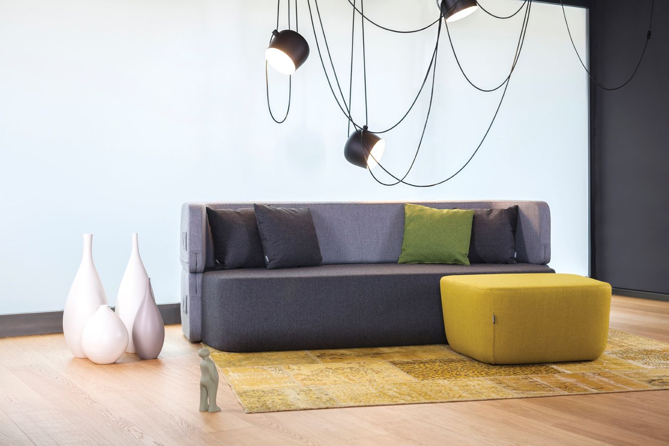 Graues Revolve Schlafsofa von Prostoria mit Kissen und gelbem Hocker auf gemustertem Teppich.