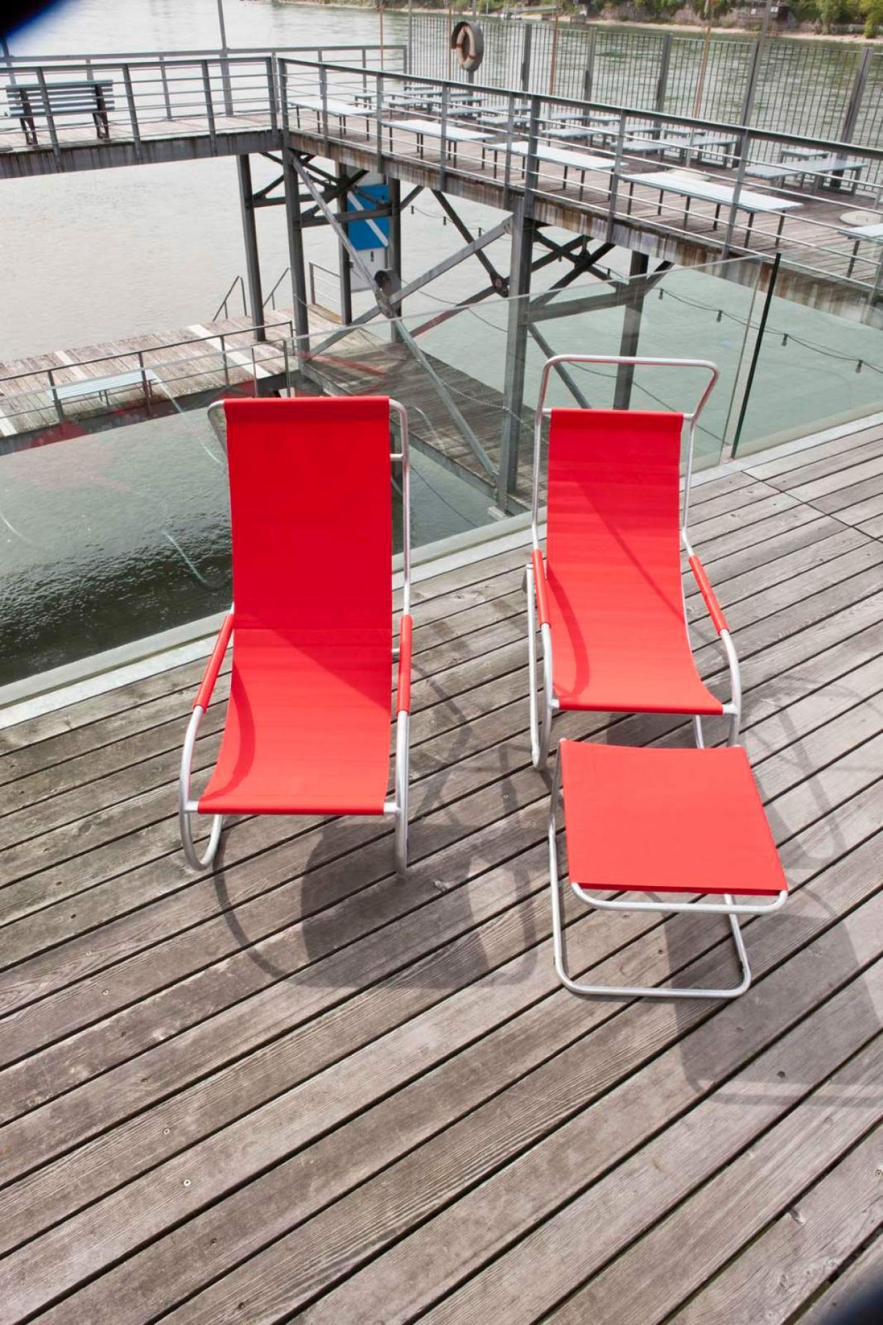 Zwei rote Lido Liegen mit Fußhocker auf Holzdeck, im Hintergrund ein Steg am Wasser.