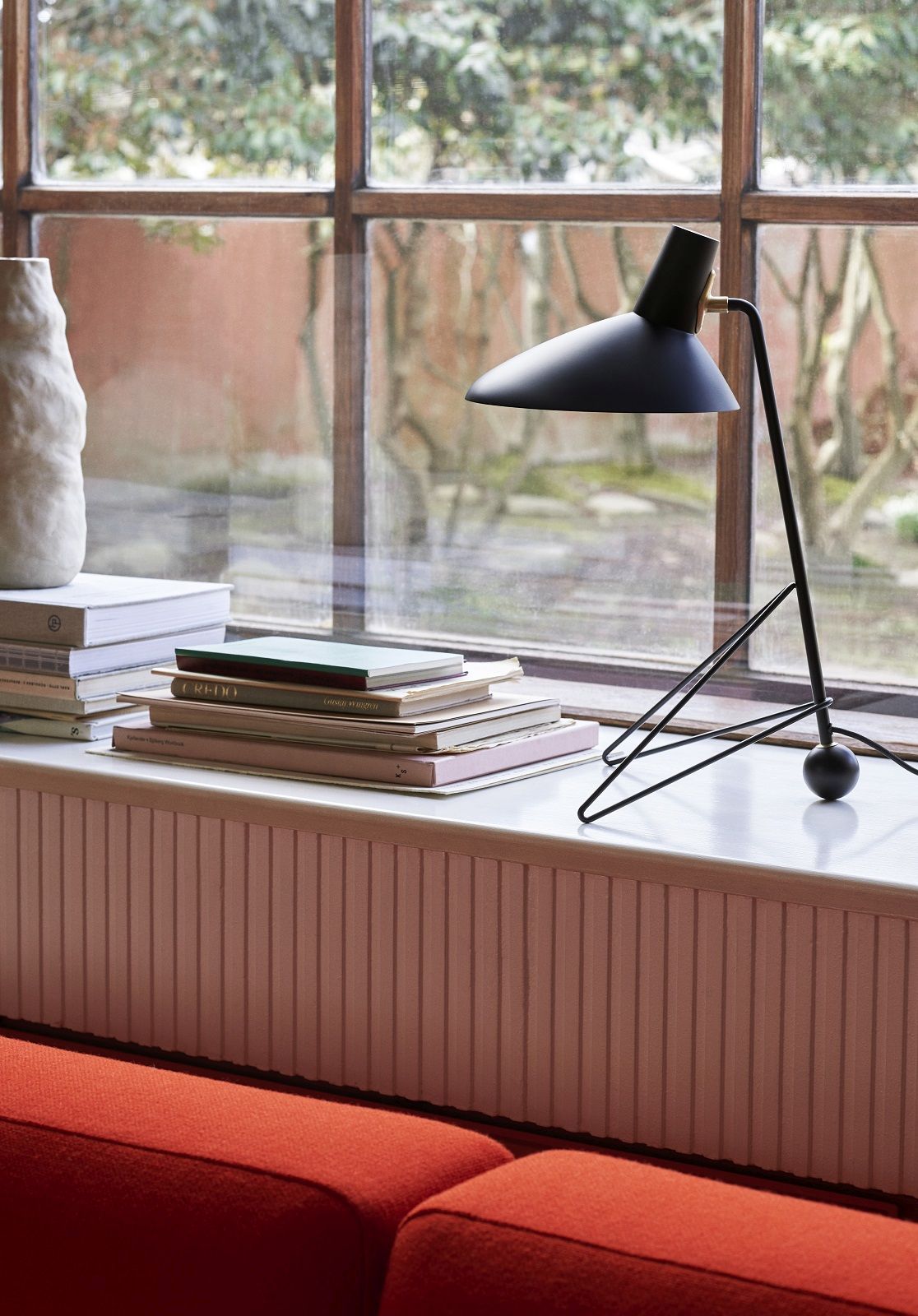 AndTradition Tripod HM9 Tischleuchte in Schwarz auf Fensterbank mit Büchern und rotem Sofa.