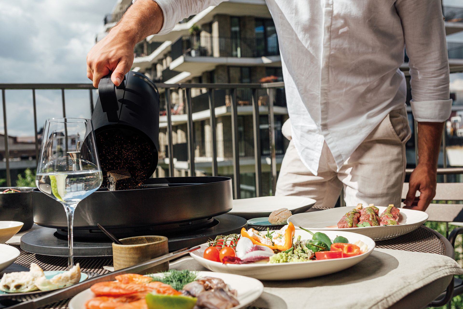 Tabl'O Tischgrill von Ofyr: Mann schüttet Holzkohle in den Grill, umgeben von Speisen auf dem Tisch.