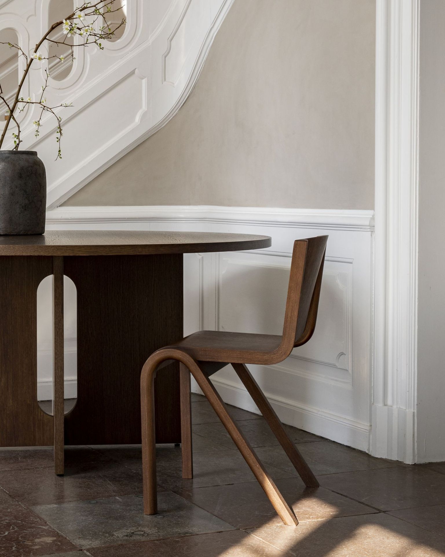 Audo Copenhagen Ready Dining Chair aus Holz neben einem dunklen Esstisch vor einer Treppe.
