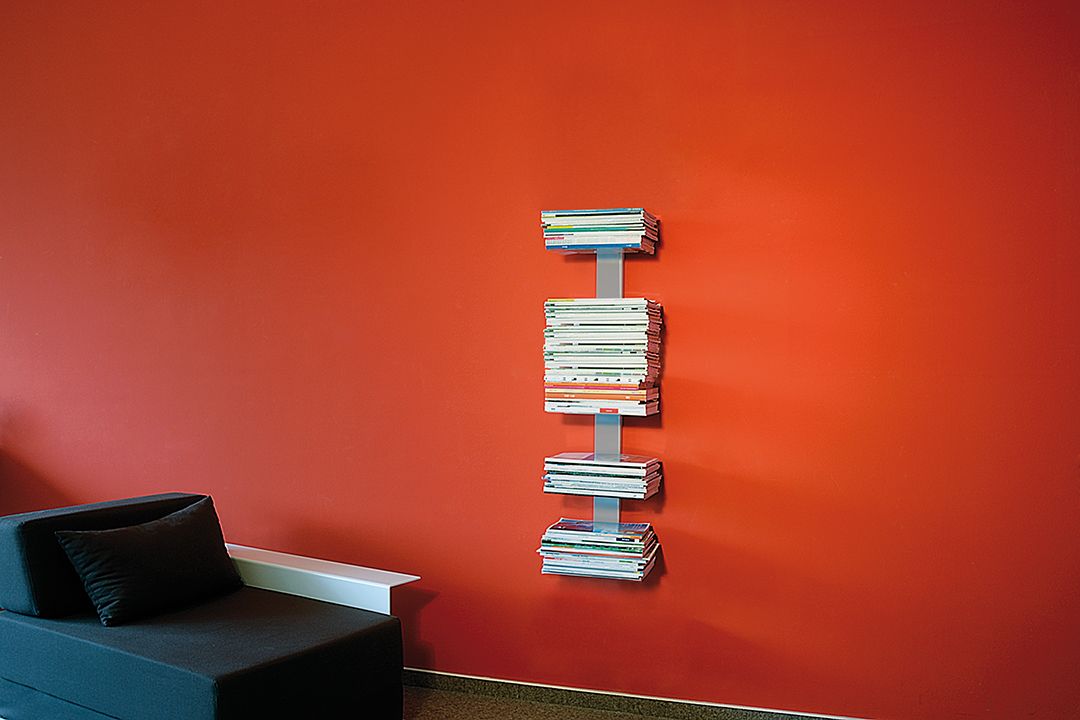 Booksbaum Wandregal mit Magazinen an roter Wand, daneben Sofa. Bücherregal für Wohnzimmer.