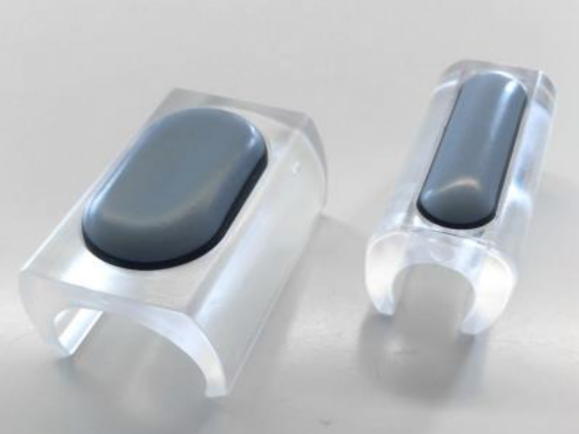 Zwei transparente Acryl-Kabelclips mit grauen Silikonpads zur Kabelführung und Organisation auf einem weißen Tisch.