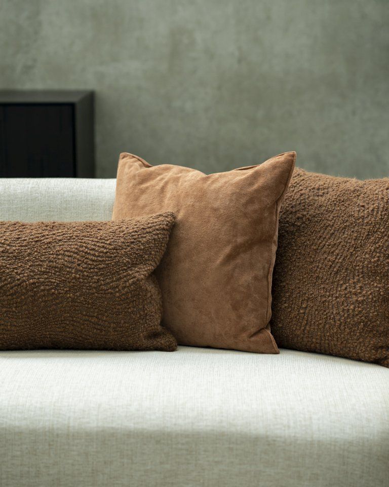 Nahaufnahme von drei braunen Kissen, darunter ein Ethnicraft Cinnamon Velvet Kissen, auf einem hellen Sofa.