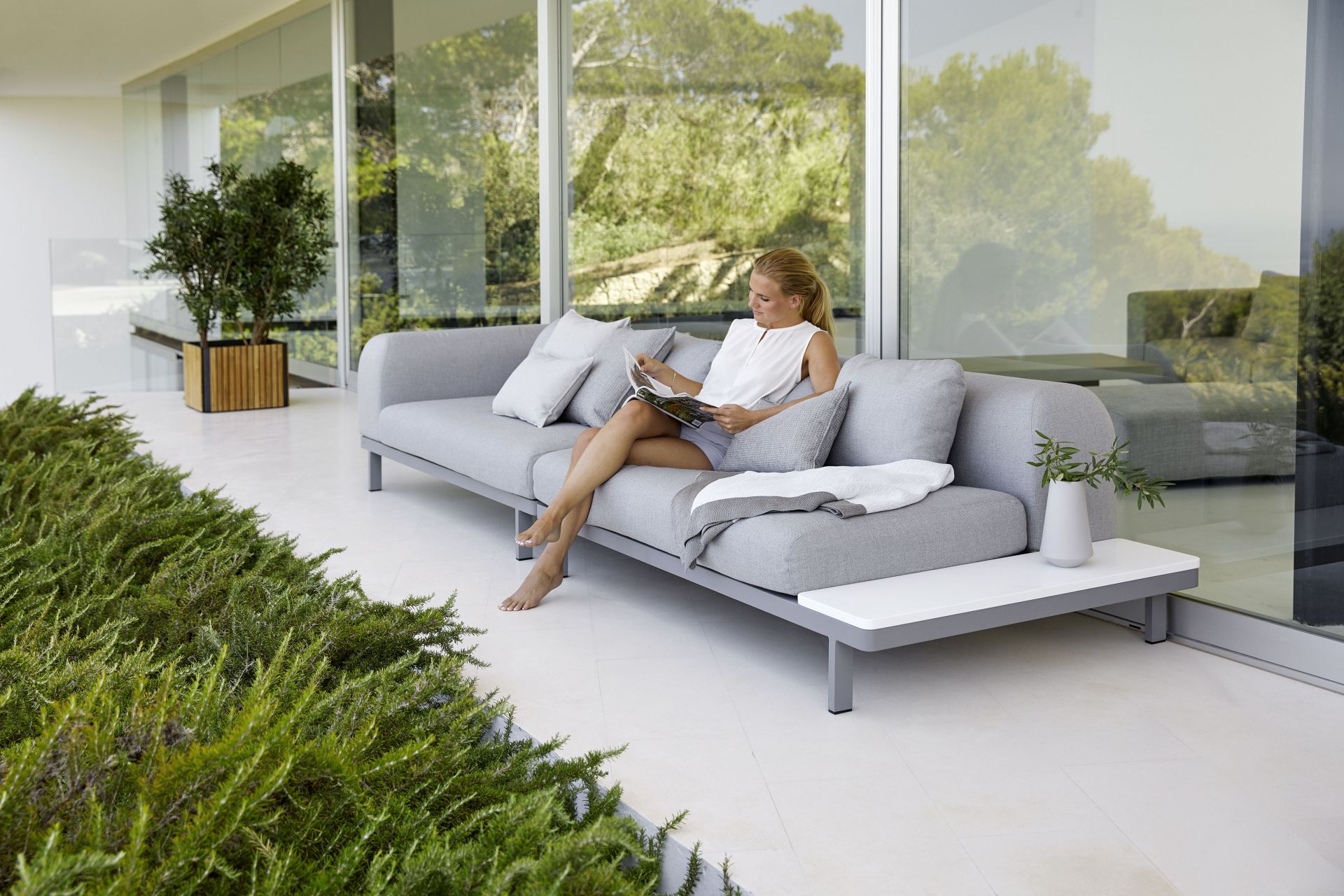 Frau liest auf einem grauen Cane-Line Sofa neben einem quadratischen Pflanzengefäß aus Holz im Garten.