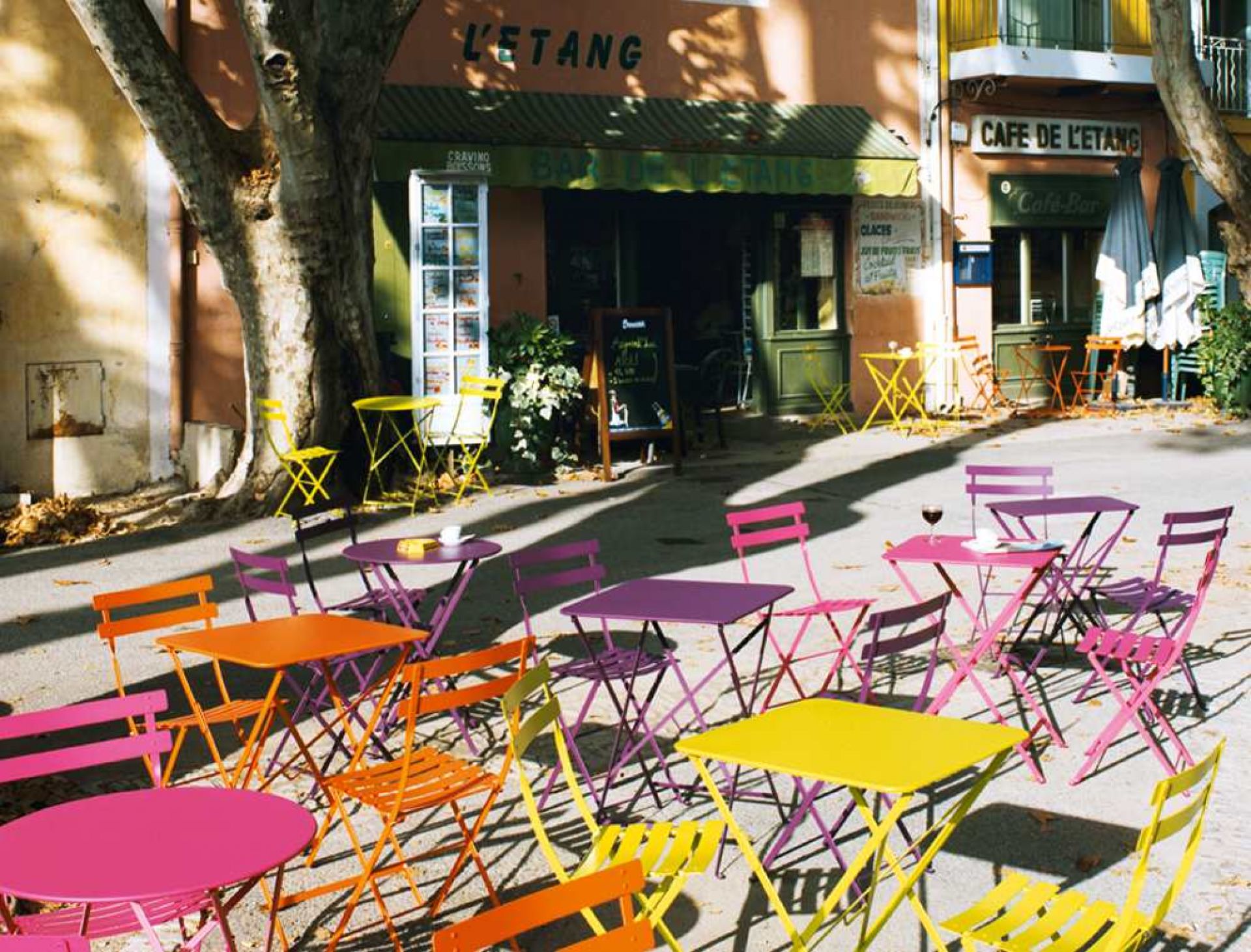 Farbige Fermob Bistro Klapptische und Stühle vor einem Café im Freien, runde und quadratische Gartentische.