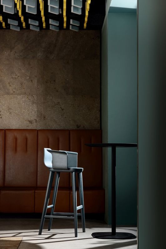 Aeon Comfort Barstool Barhocker mit Armlehnen in grau vor brauner Wand und Stehtisch.
