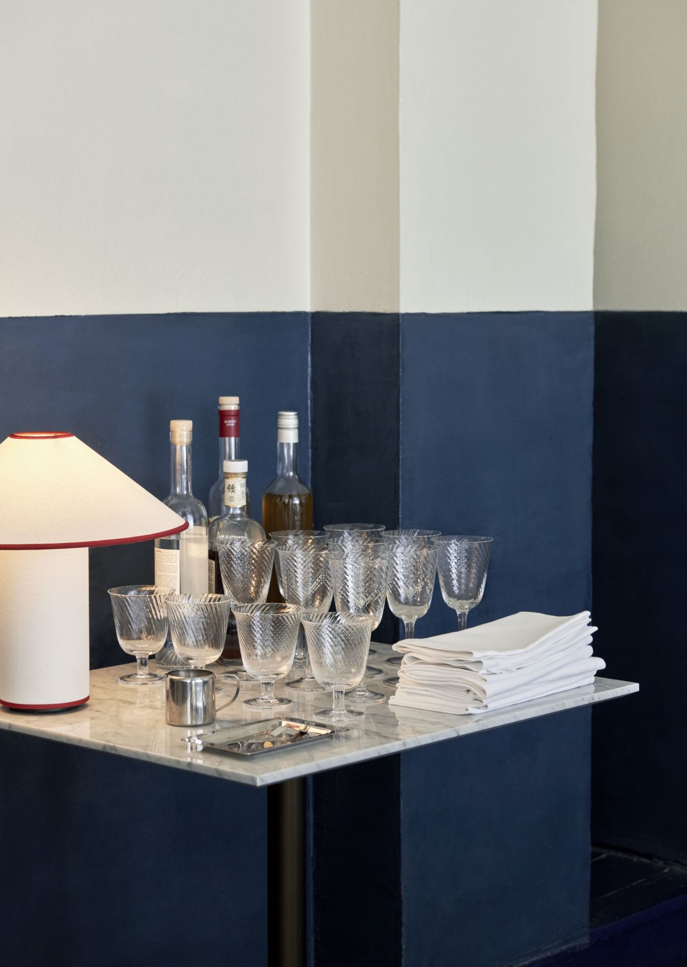 AndTradition Colette ATD6 Tischleuchte auf Marmortisch mit Gläsern, Karaffen und Servietten vor blau-weißer Wand.