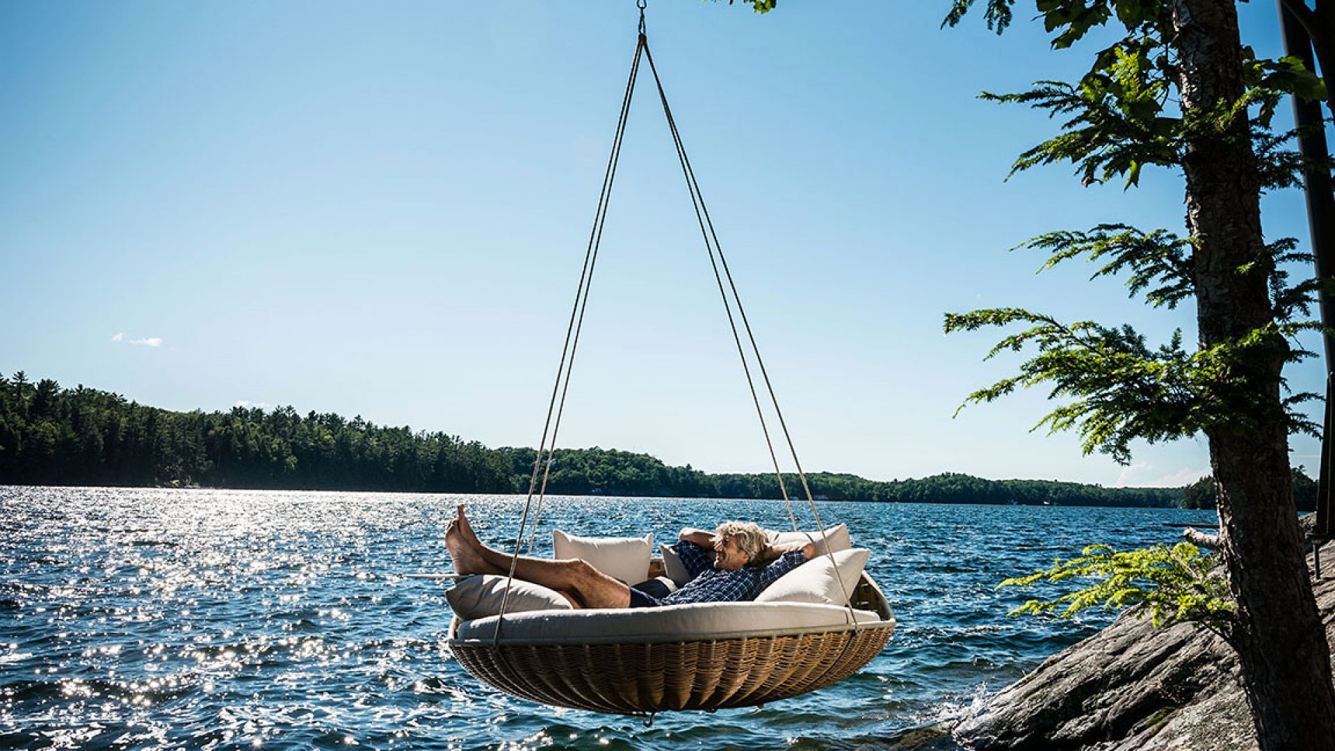 Mann entspannt in Dedon Swingrest Hängelounge mit Rückenkissen am See. Outdoor-Möbel für Garten und Terrasse.