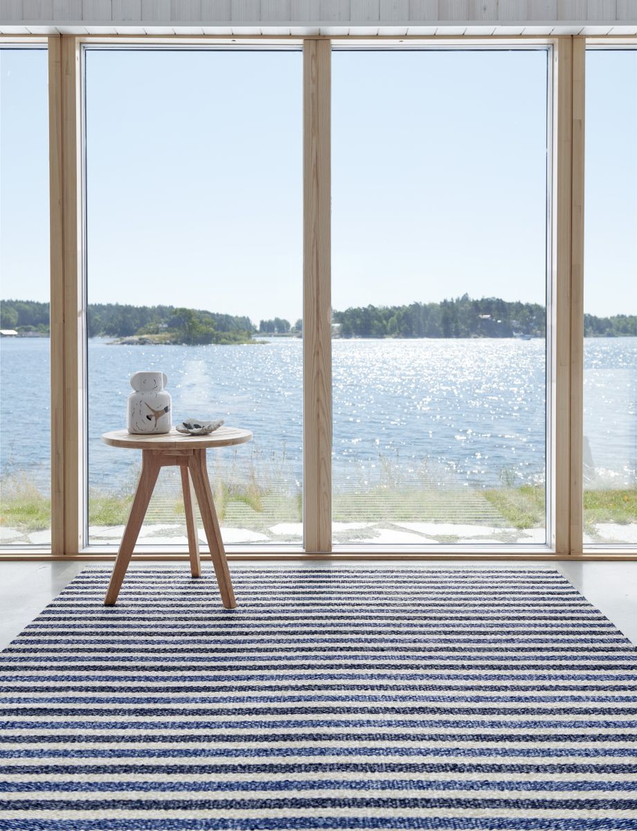 Pappelina Teo Teppich 230x320 cm, blau-weiß gestreift, vor Fenster mit Meerblick und kleinem Tisch.