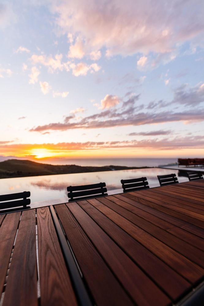 Alua Armrest Dining Chair: Elegante Outdoor Armlehnstühle aus Holz an einem Tisch mit Meerblick bei Sonnenuntergang.