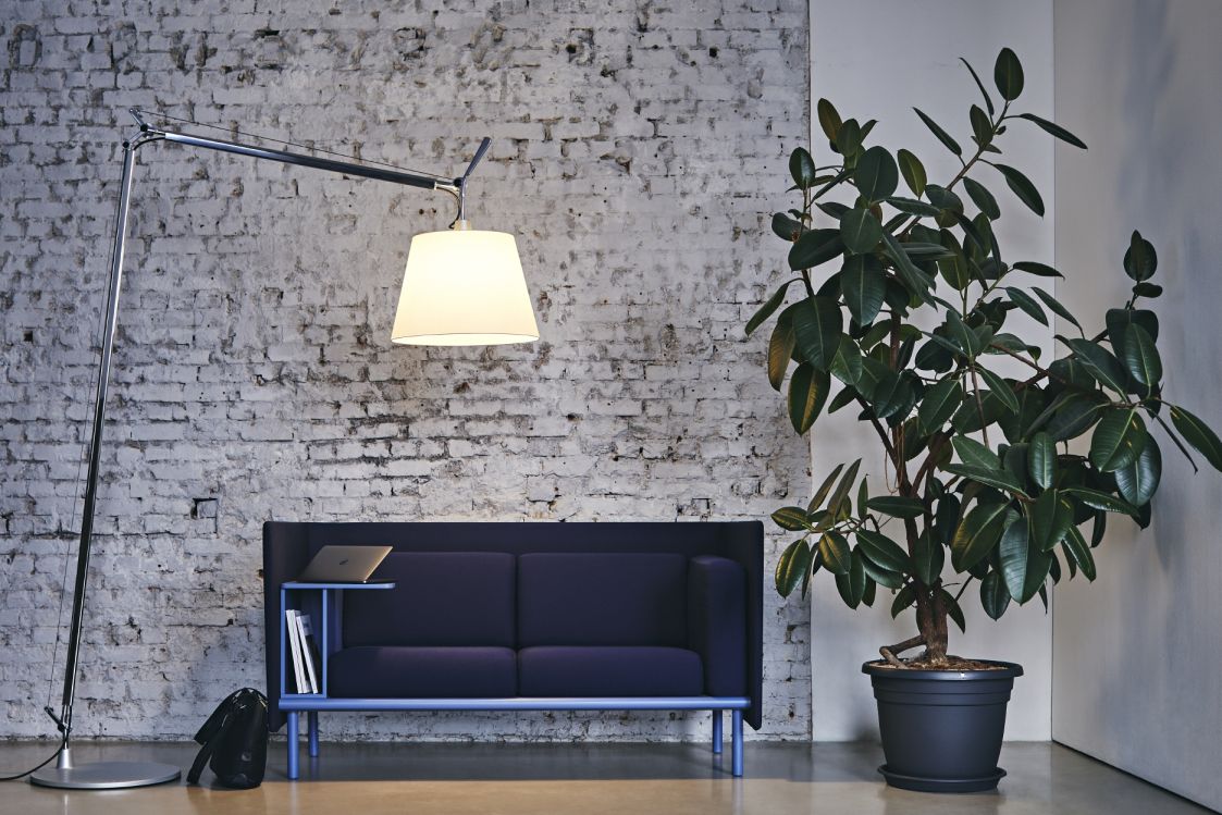 Floater 2-Sitzer Sofa von Cor in blau mit Stehleuchte und Zimmerpflanze vor Backsteinwand.