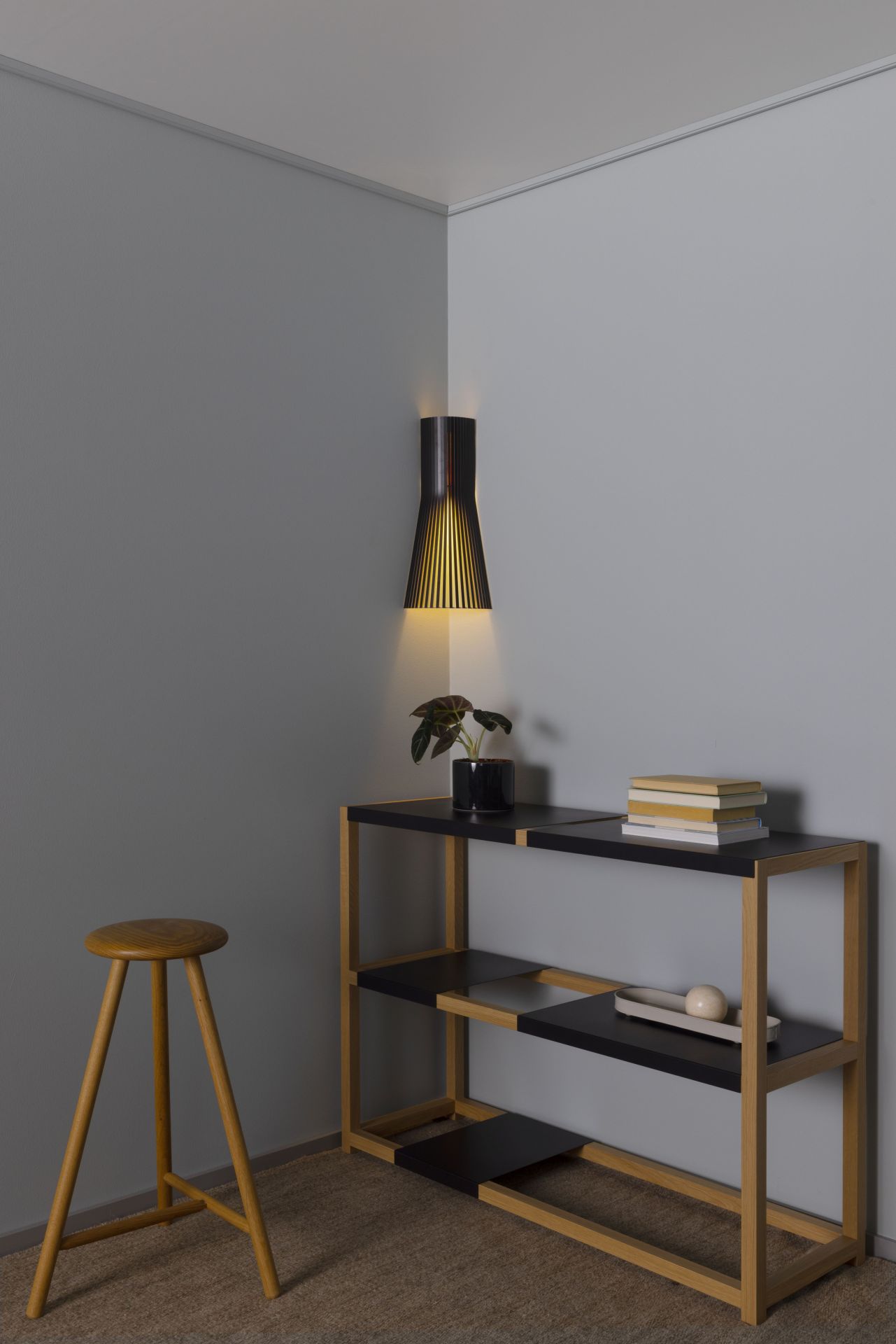Secto 4237 Eckleuchte aus Walnussfurnier an grauer Wand, neben Regal und Hocker.