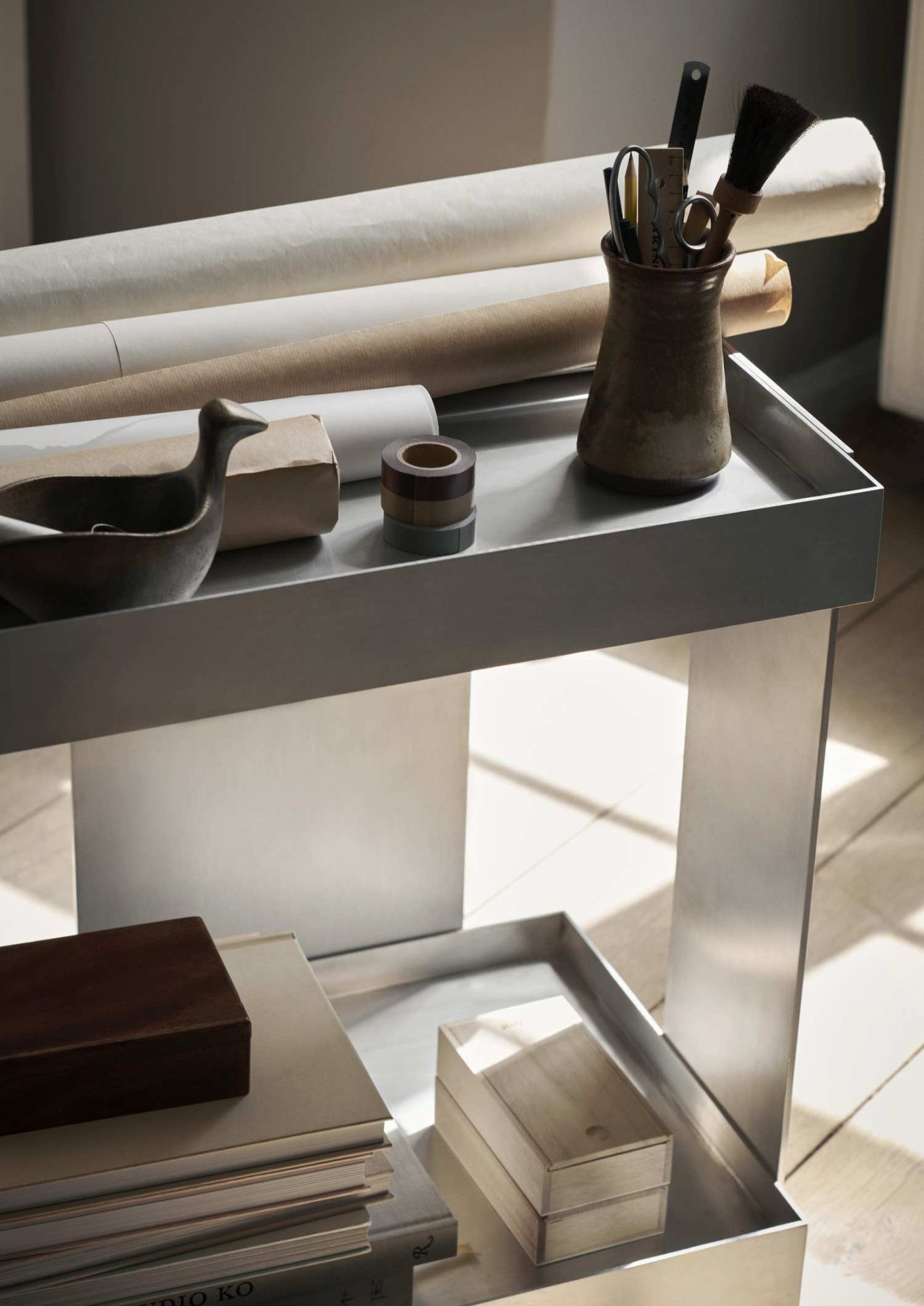 Ferm Living Lager Trolley: Moderner Aluminium Servierwagen mit zwei Ebenen, dekoriert mit Büroartikeln und Papieren.
