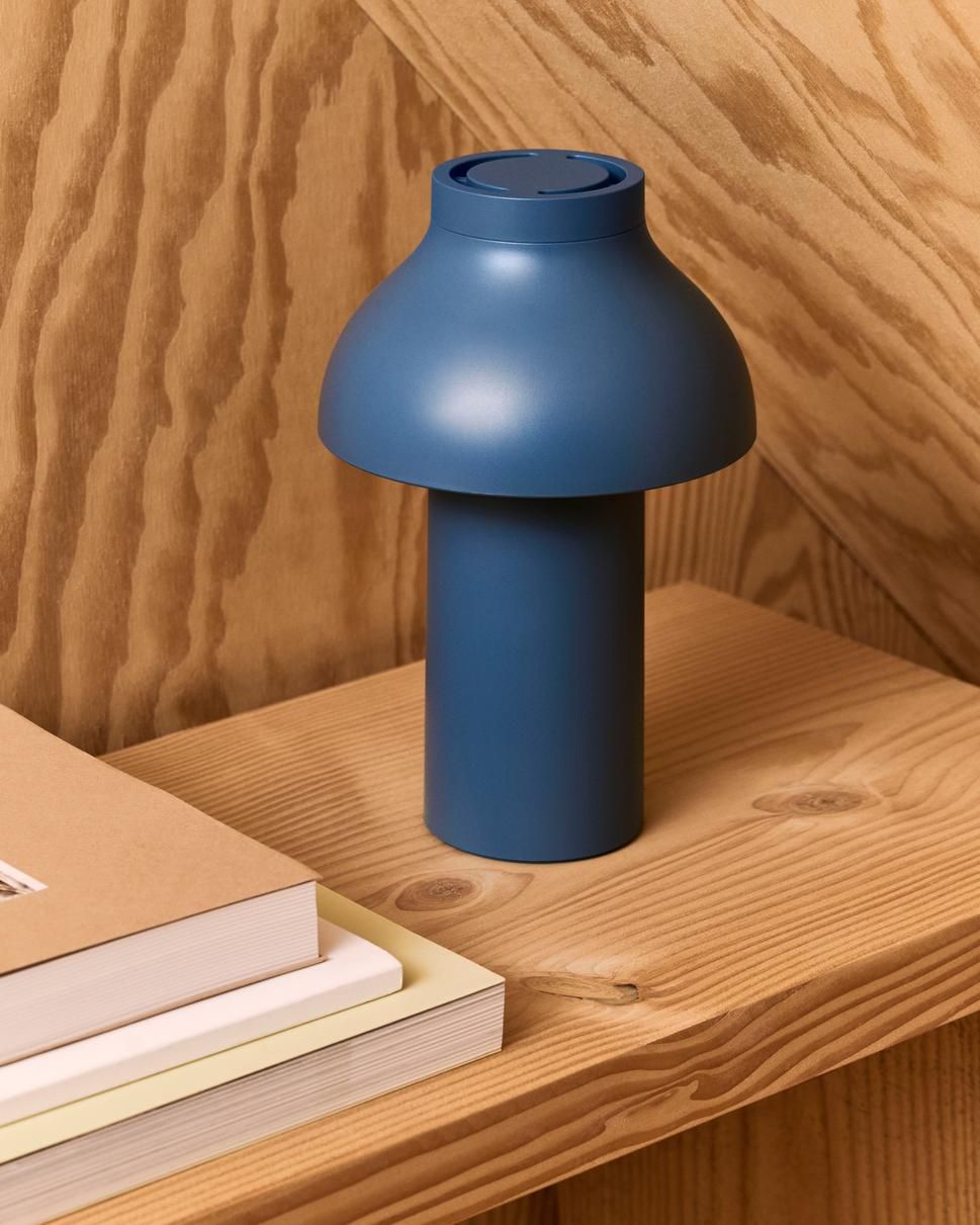 Blaue PC Portable Akkuleuchte von Hay auf einem Holztisch, daneben Bücher gestapelt. Moderne Tischleuchte.