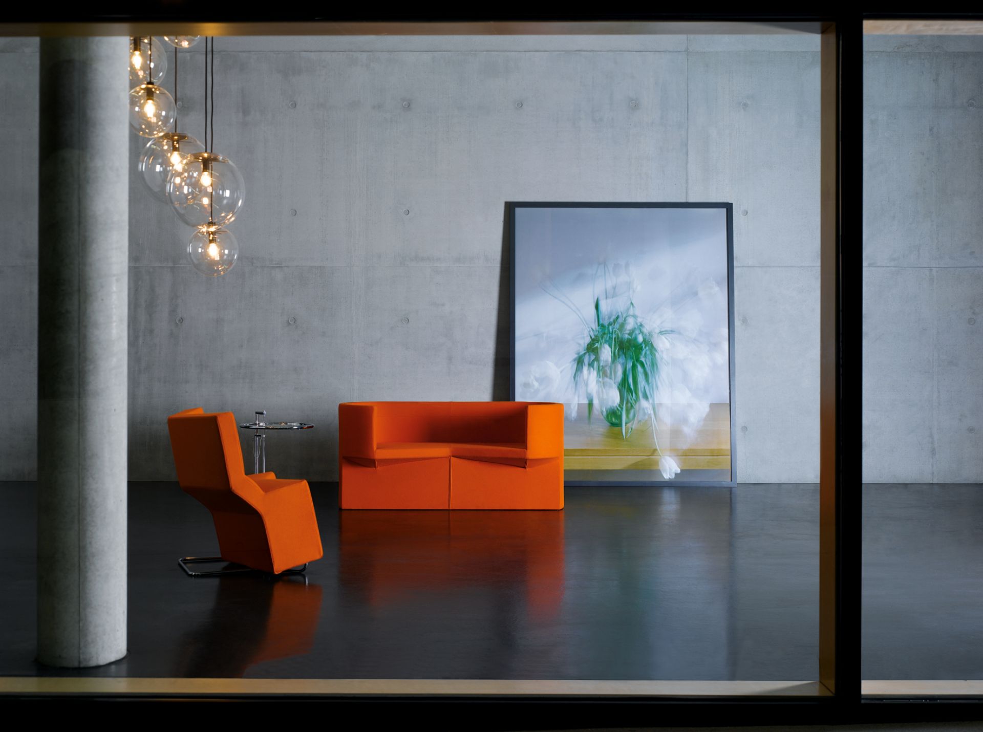 Moderner Wohnraum mit orangefarbenem Sessel, Sofa und ClassiCon E1027 Beistelltisch mit Glasplatte vor Betonwand.