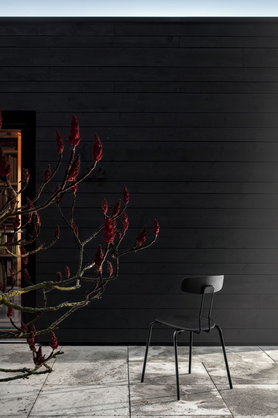 Schwarzer Okito Chair Stuhl Zeitraum vor dunkler Holzwand und blühendem Strauch, modernes Möbeldesign.