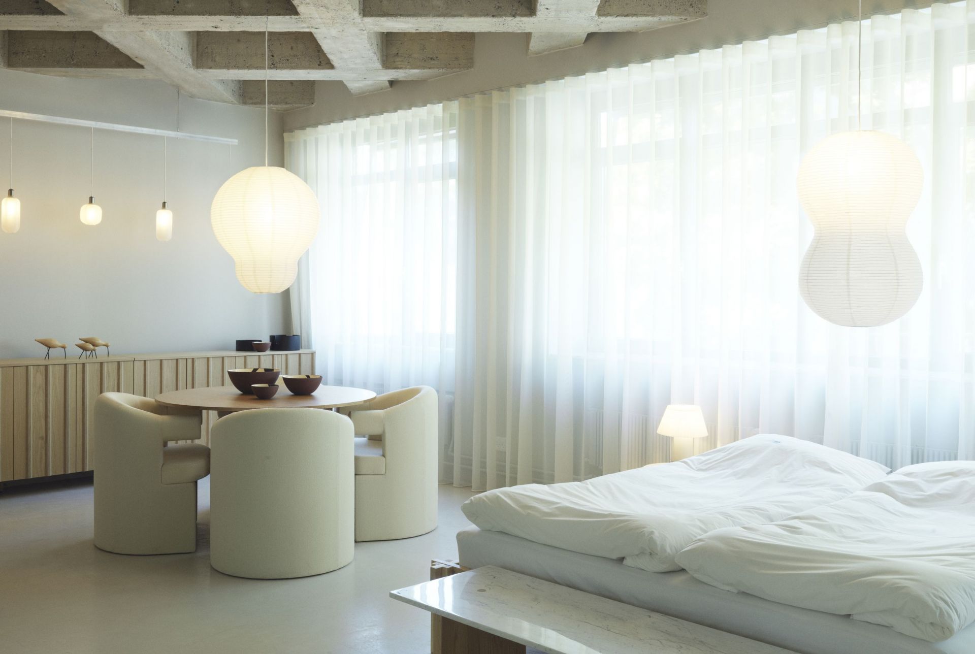 Helles Interieur mit Normann Copenhagen Burra Stuhl, Holztisch, Lampen und Bett mit weißer Bettwäsche.