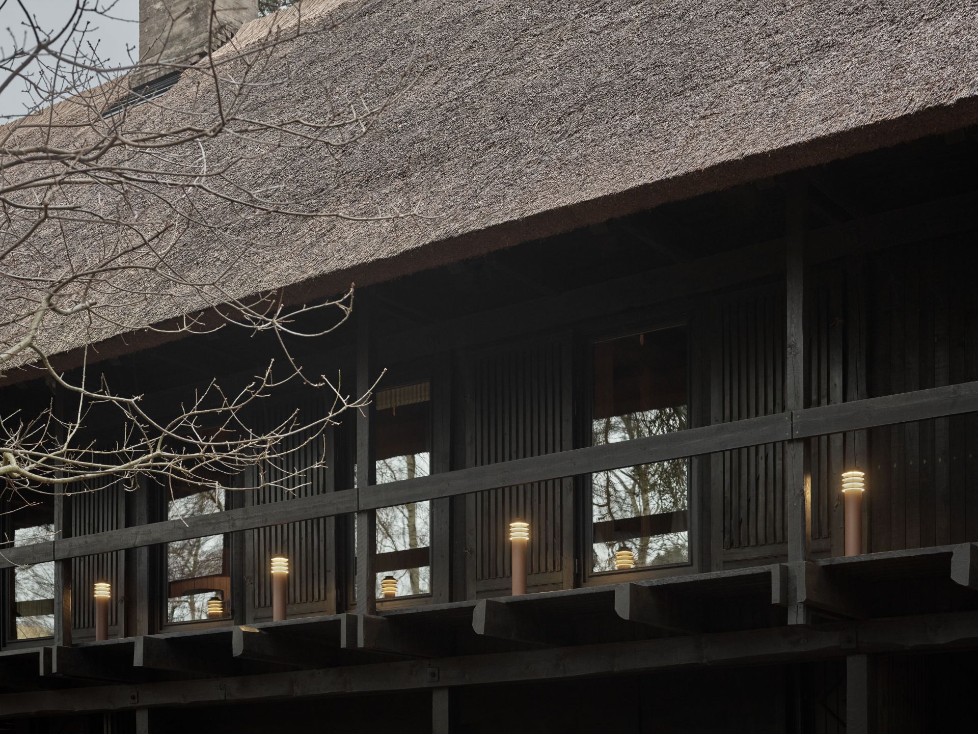 Nahaufnahme der Louis Poulsen Bysted Garden Pollerleuchten vor einem dunklen Holzhaus mit Reetdach.