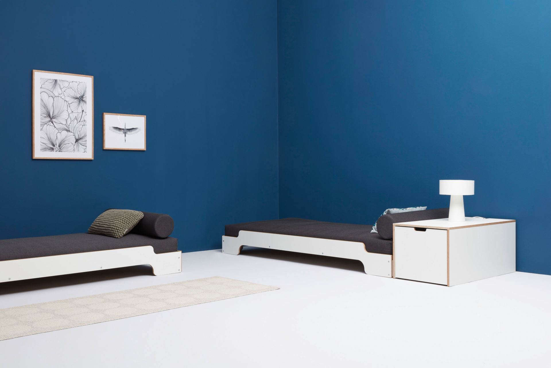 Zwei weiße Stapelliegen von Müller Möbelwerkstätten mit grauen Auflagen und Kissen vor blauer Wand.