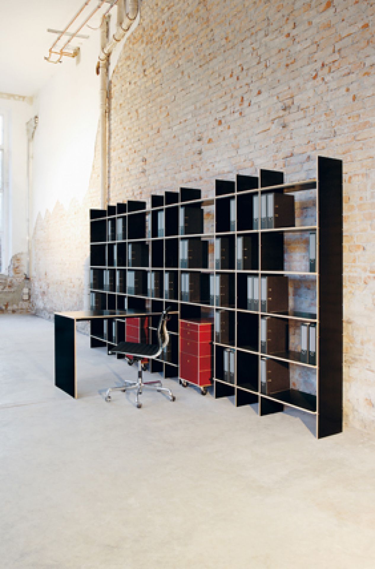 Schwarzes FNP Regal von Nils Holger Moormann mit Schreibtisch und roten Rollcontainern vor Backsteinwand.