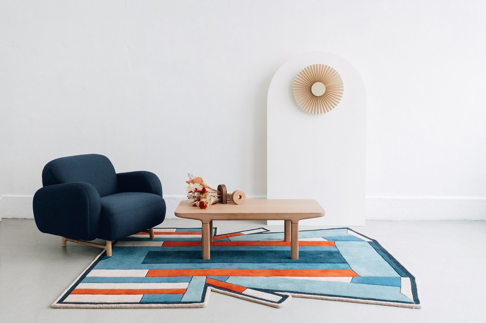 Wohnzimmer-Szene mit blauem Sessel, Holztisch, geometrischem Teppich und Carmen Wall Lampe Wandleuchte als Dekoration.