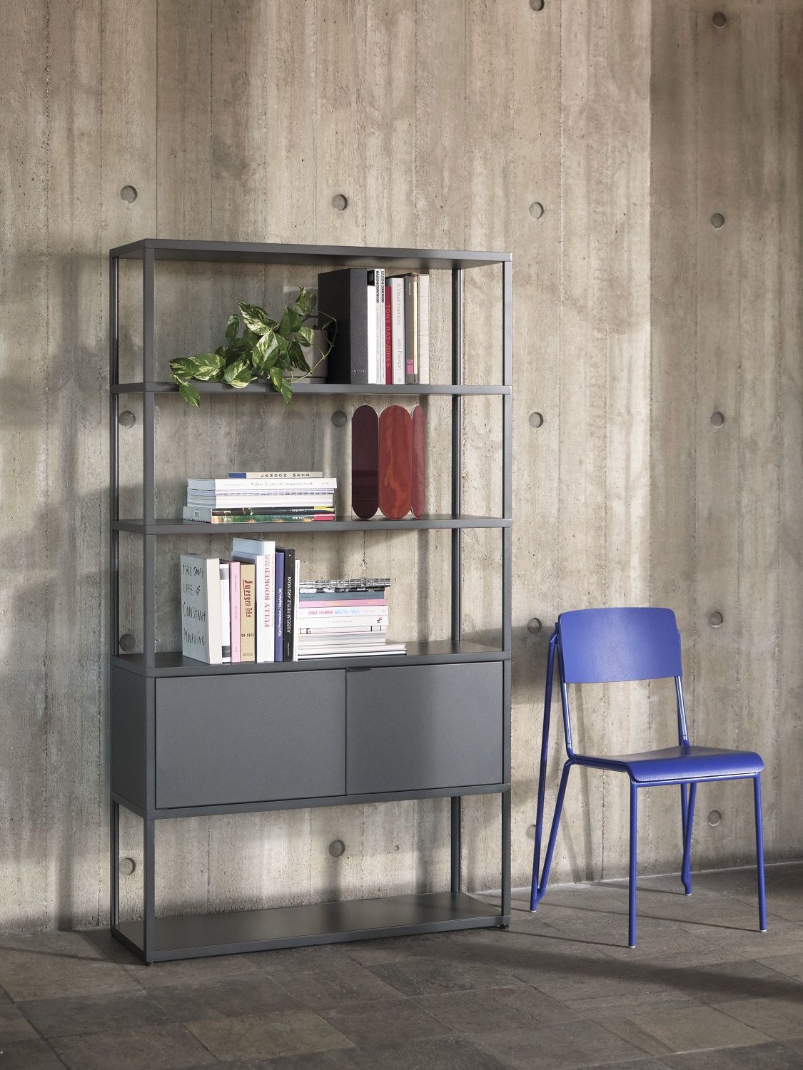 Graues New Order Sideboard von Hay mit Regalen, Schrankfächern und blauer Stuhl vor Betonwand.