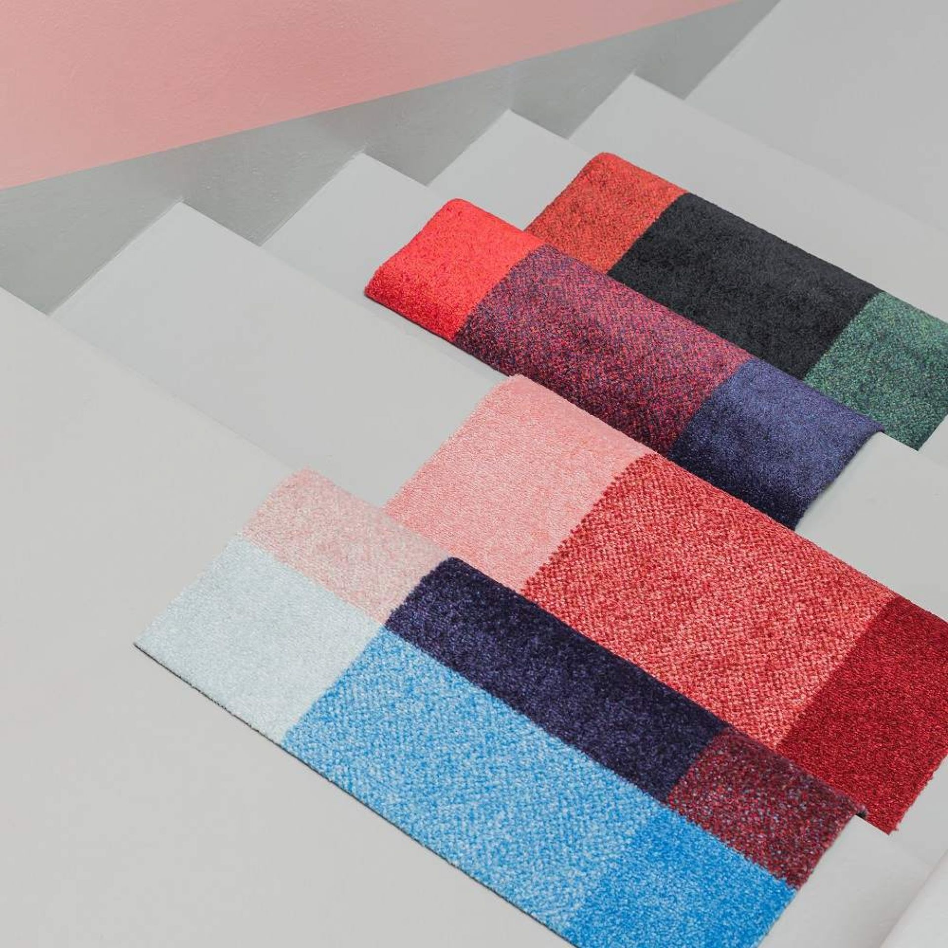 Mehrere farbenfrohe Heymat Fußmatten für Innen und Außen auf einer grauen Treppe, verschiedene Designs.