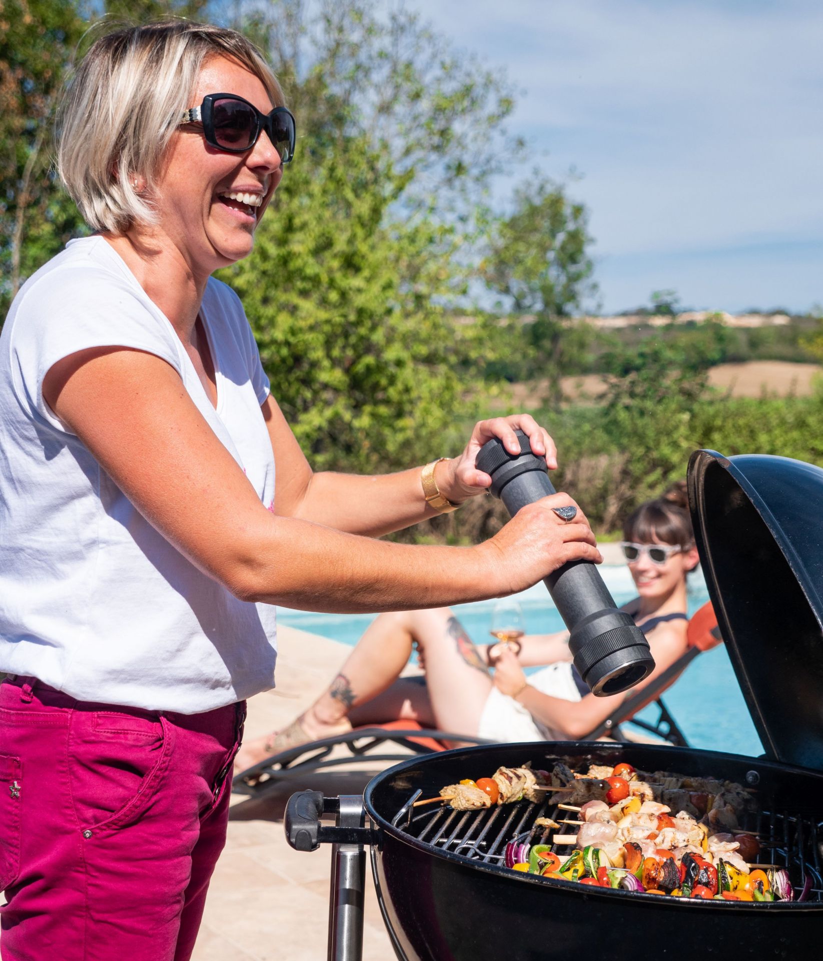 Frau würzt Grillspieße mit Peugeot BBQ Pfeffermühle, während Freundin am Pool entspannt.