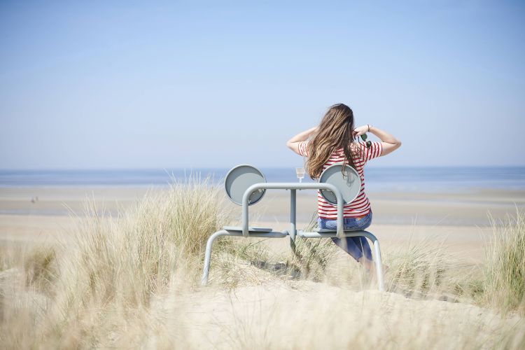 Frau sitzt auf Extremis Bistroo Stuhl-Tisch-Kombination am Strand mit Meerblick, Outdoor Möbel für Gastronomie und Garten.
