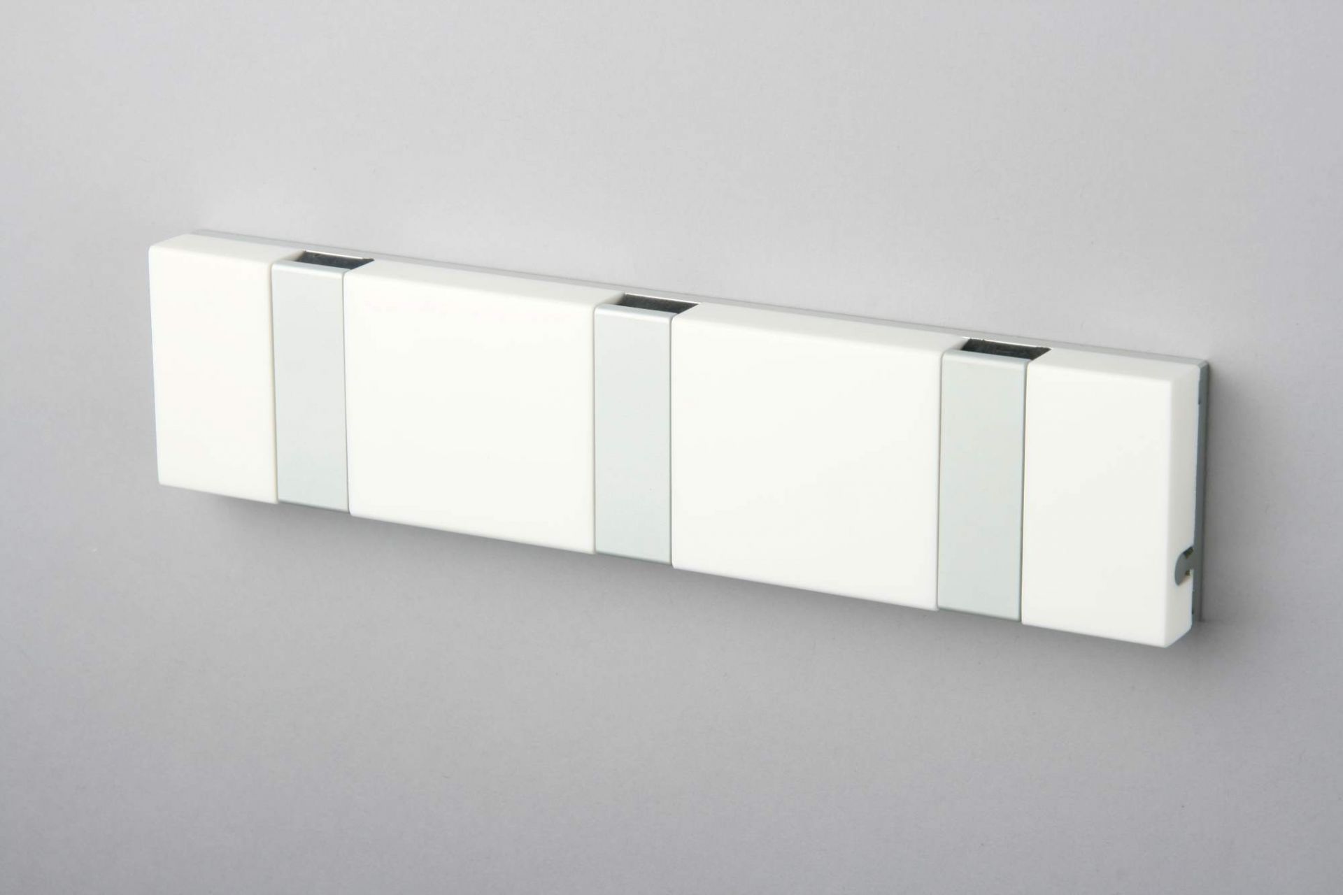 Weiße Knax Lite Wandgarderobe mit drei Haken, waagerecht an grauer Wand montiert. Moderne Garderobenleiste für Flur und Diele.