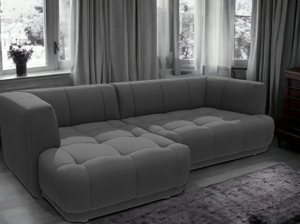 Graues Quilton Sofa von Hay mit Ottomane und gesteppten Polstern vor Fenster mit Vorhängen.
