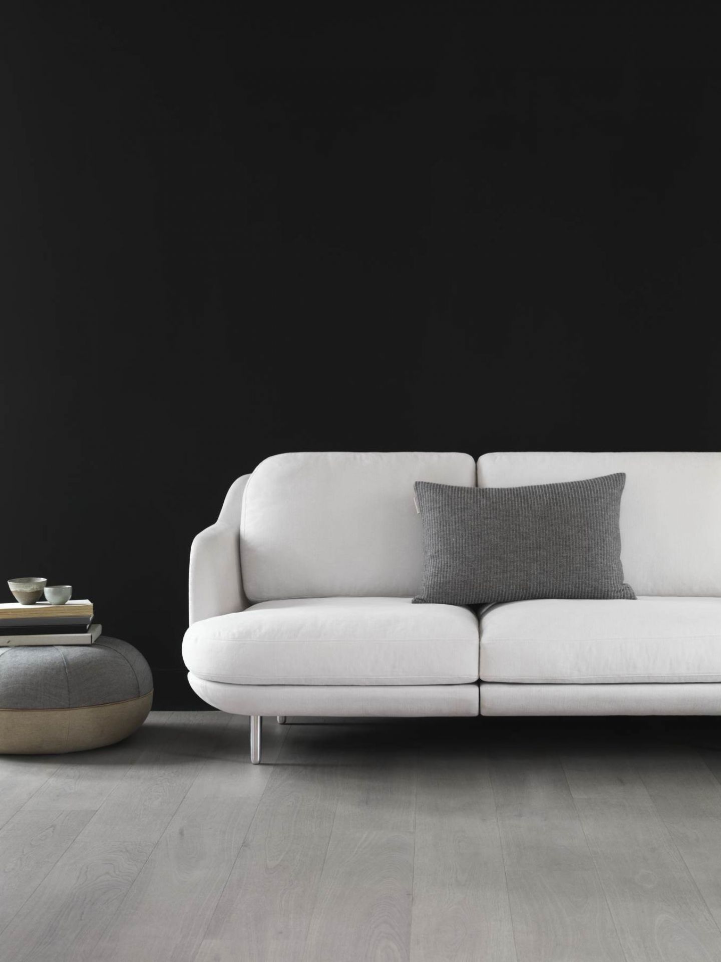 Helles Lune Sofa von Fritz Hansen mit grauem Kissen und Hocker vor schwarzer Wand.