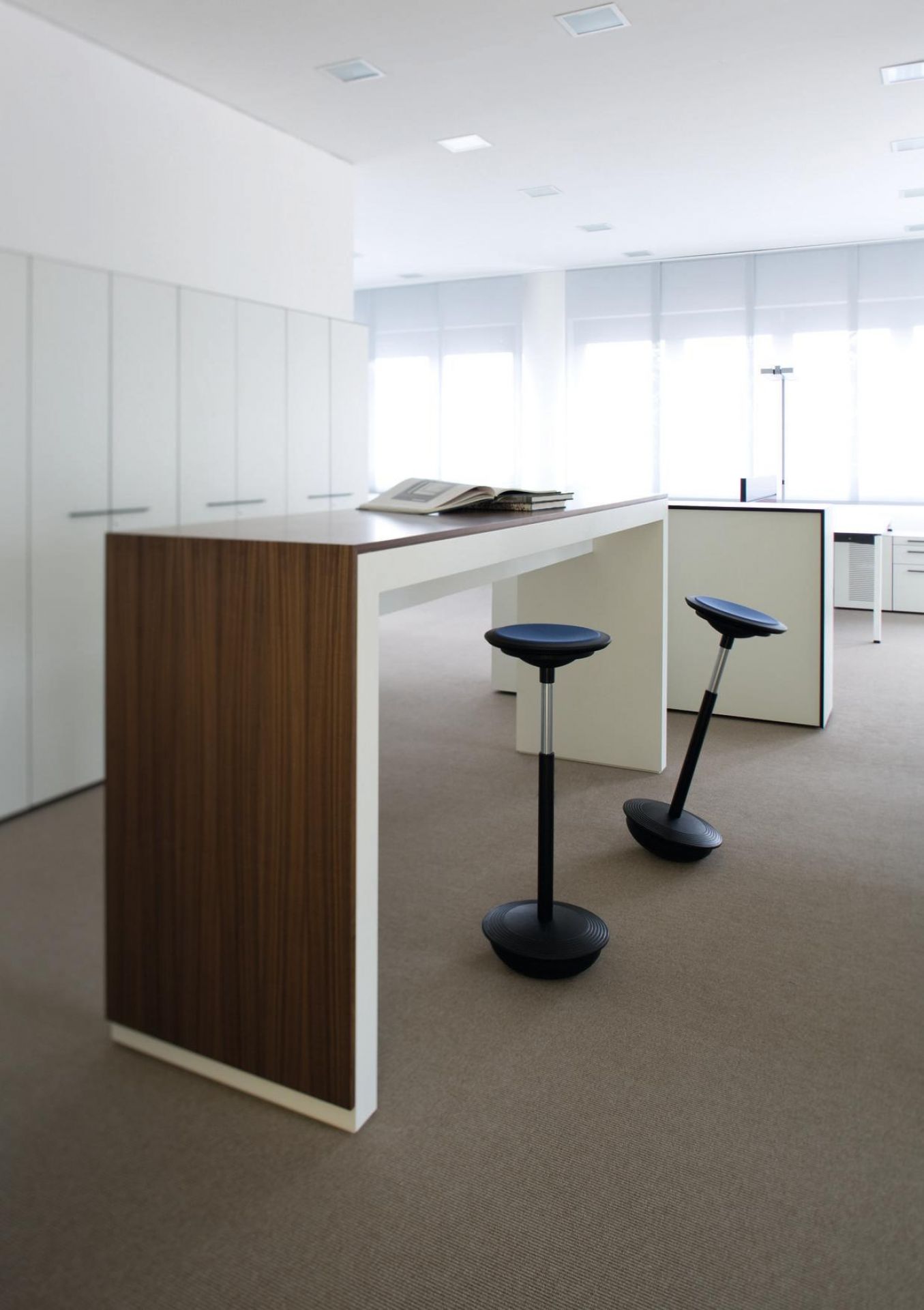 Zwei Wilkhahn Stitz Sitz- / Stehhocker in einem hellen Büro mit Stehtisch und Teppichboden.