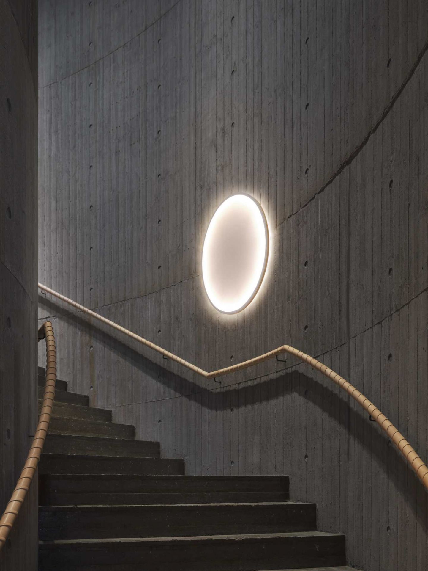 Ovale Calm Wall Lamp Wandleuchte von Muuto an einer Betonwand neben einer Treppe.
