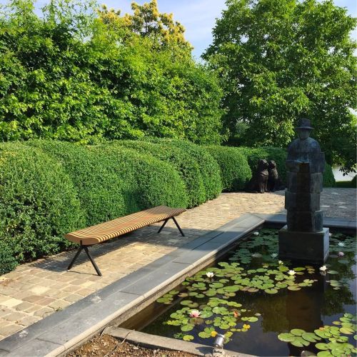 Prelude Bank: Moderne Holzbank im Park neben einem Teich mit Seerosen.