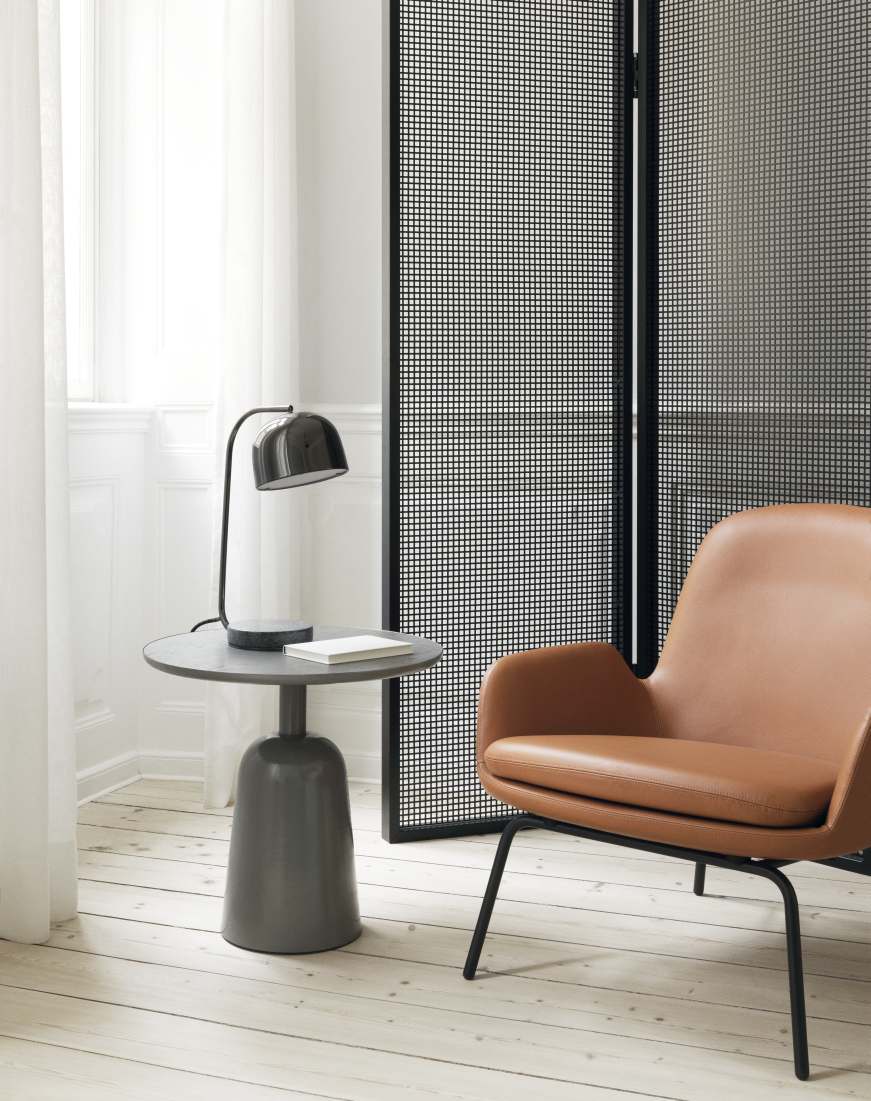 Grauer Normann Copenhagen Turn Table Beistelltisch mit Lampe und Buch neben einem Sessel.