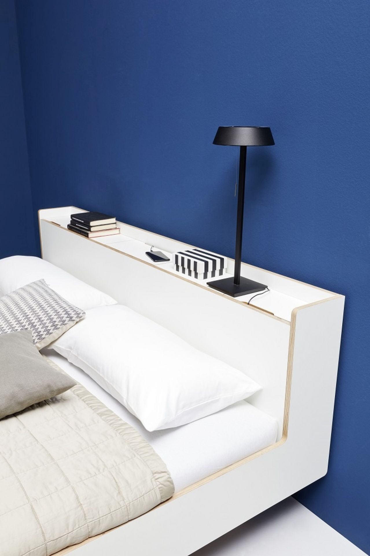 Nook Einzelbett von Müller Möbelwerkstätten in Weiß mit Ablagefläche und Nachttischlampe vor blauer Wand.