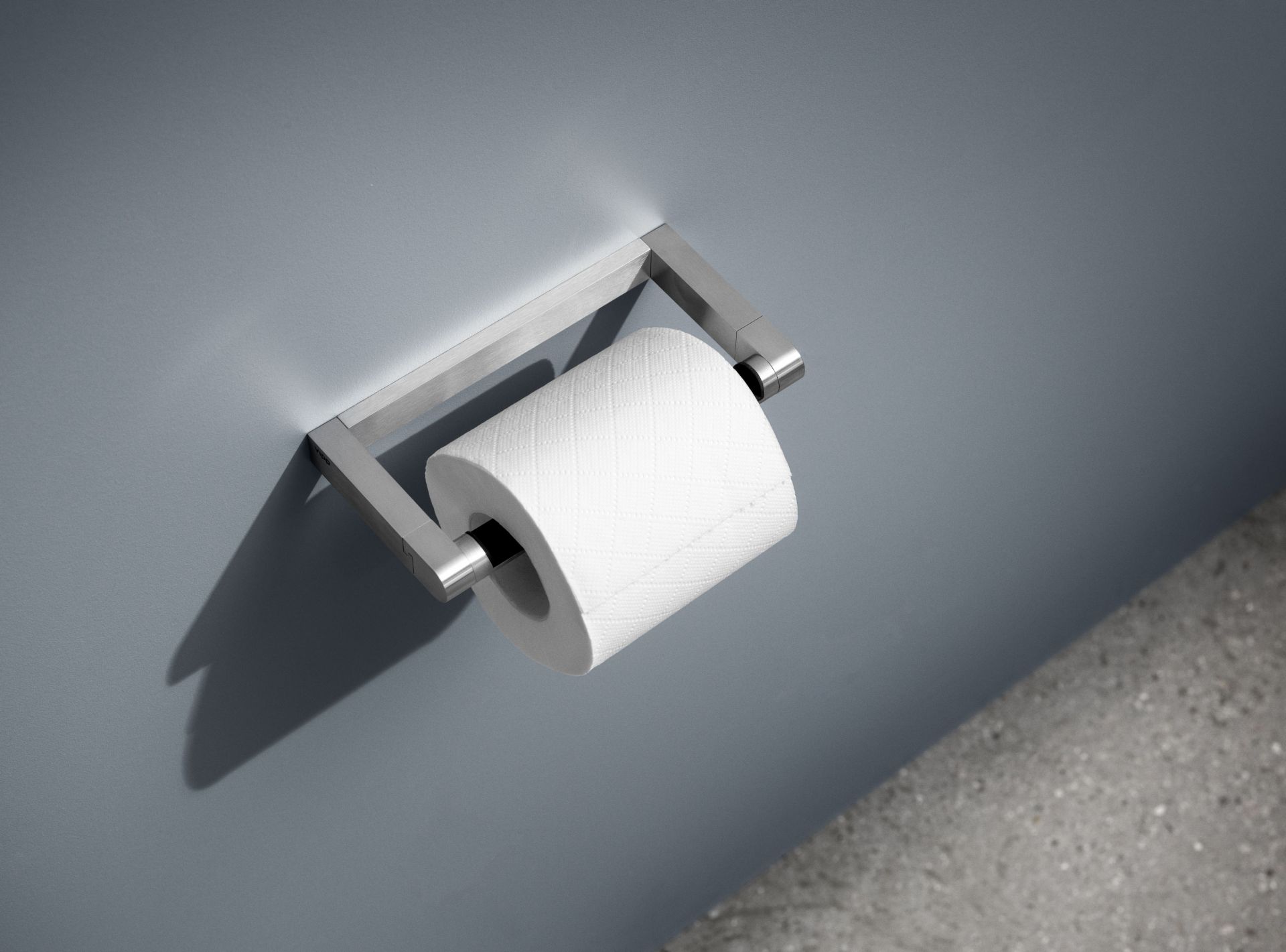 Vipp WC-Rollenhalter aus gebürstetem Edelstahl mit einer weißen Toilettenpapierrolle an grauer Wand.