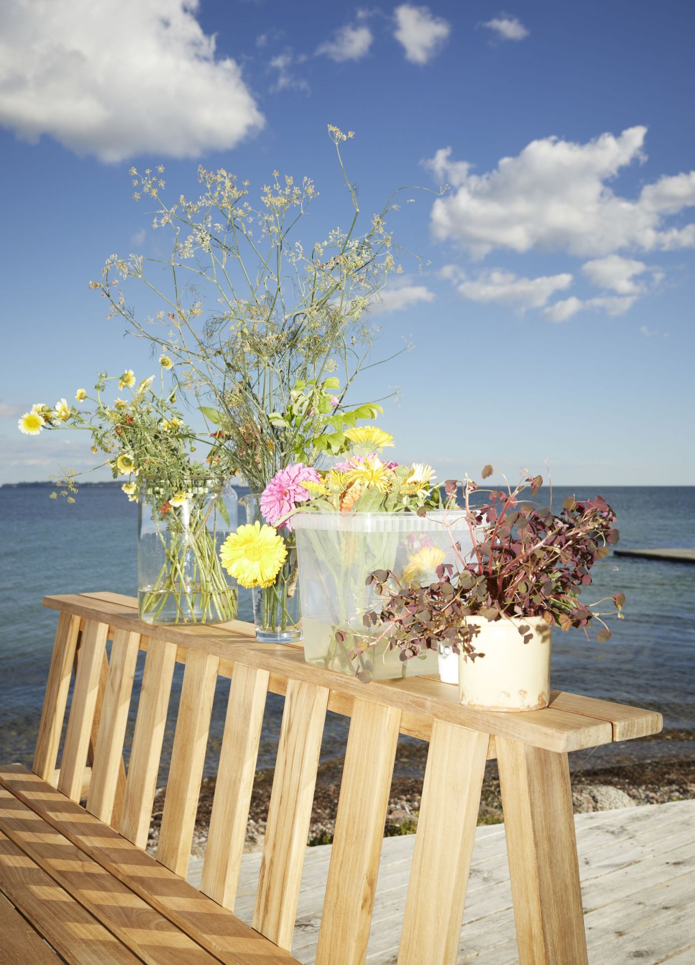 Cadence Bank Teak Outdoor Bank mit Blumenvasen und Topfpflanze vor blauem Himmel und Meer.
